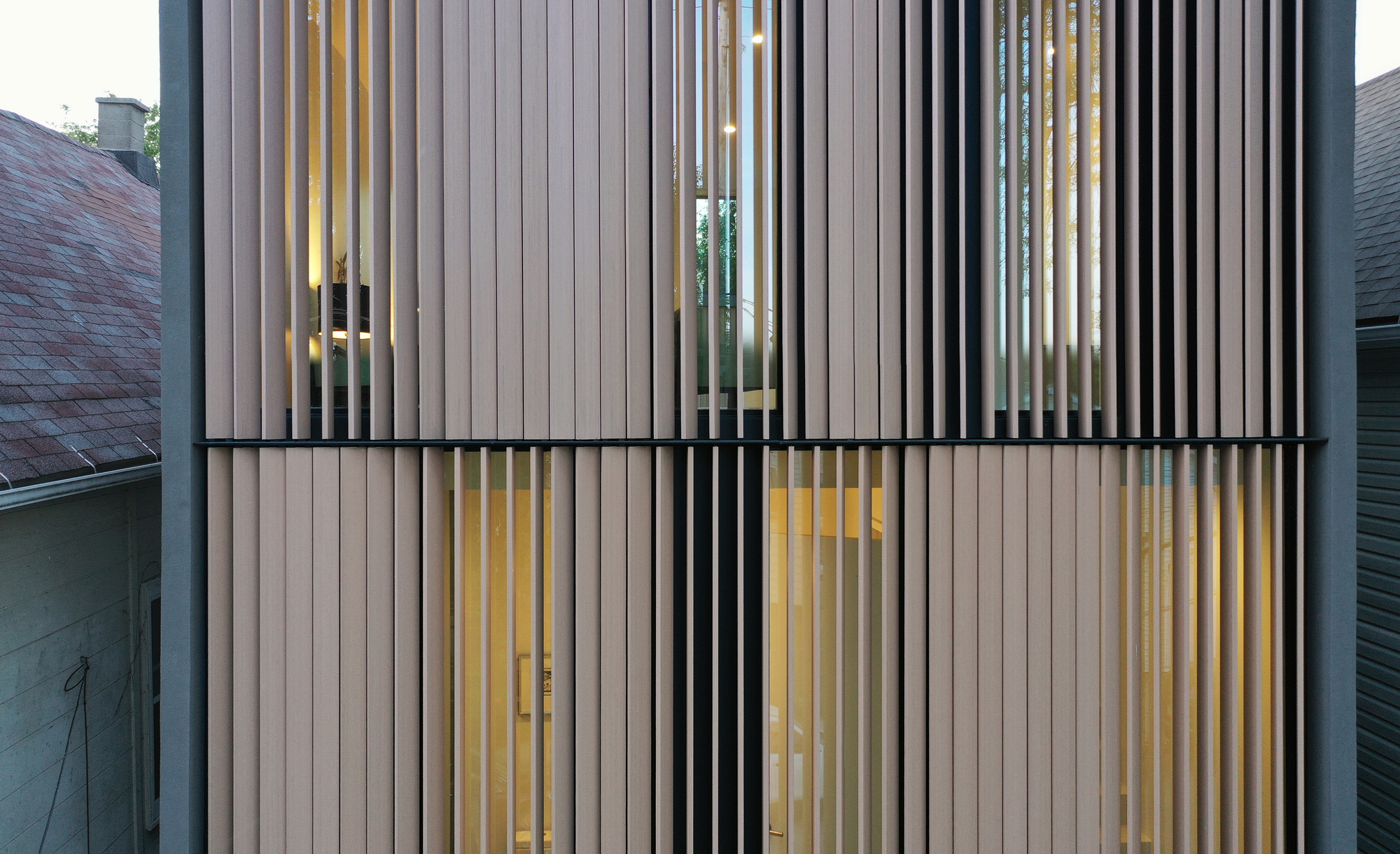 Close view of the vertical timber fin screen over a two-storey facade, glowing with warm interior light at dusk