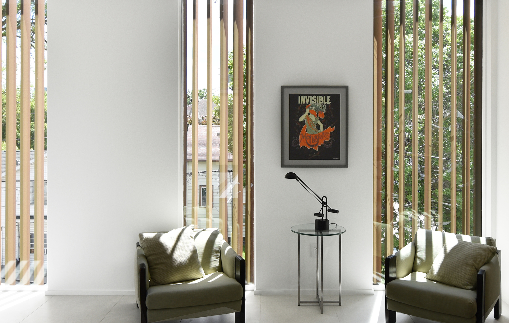 Sitting corner with two olive armchairs and a glass side table, framed by vertical timber fins and leafy trees outside