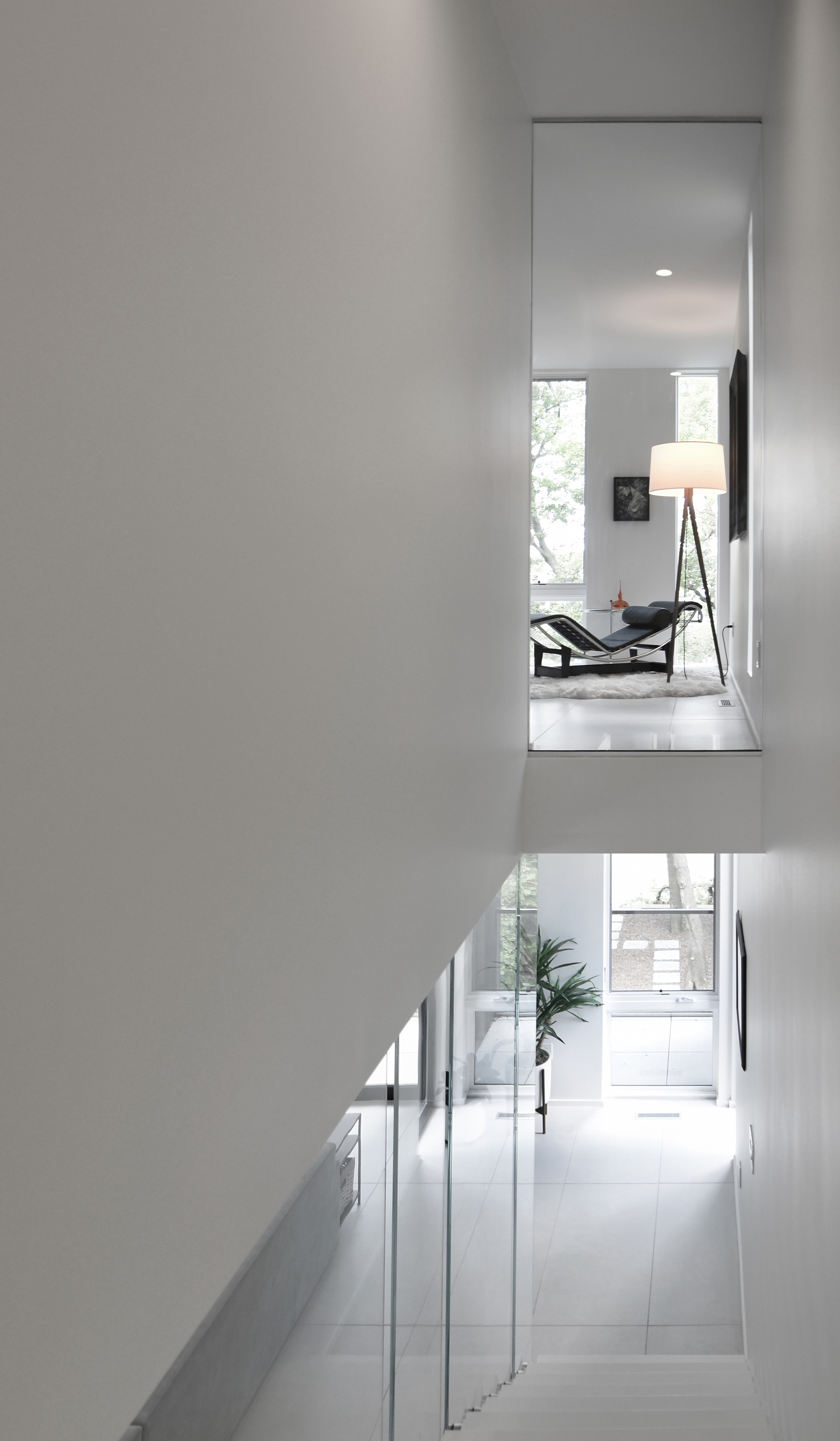 Stairwell view looking up past a white wall to a lounge chair and tripod floor lamp, with a potted palm below