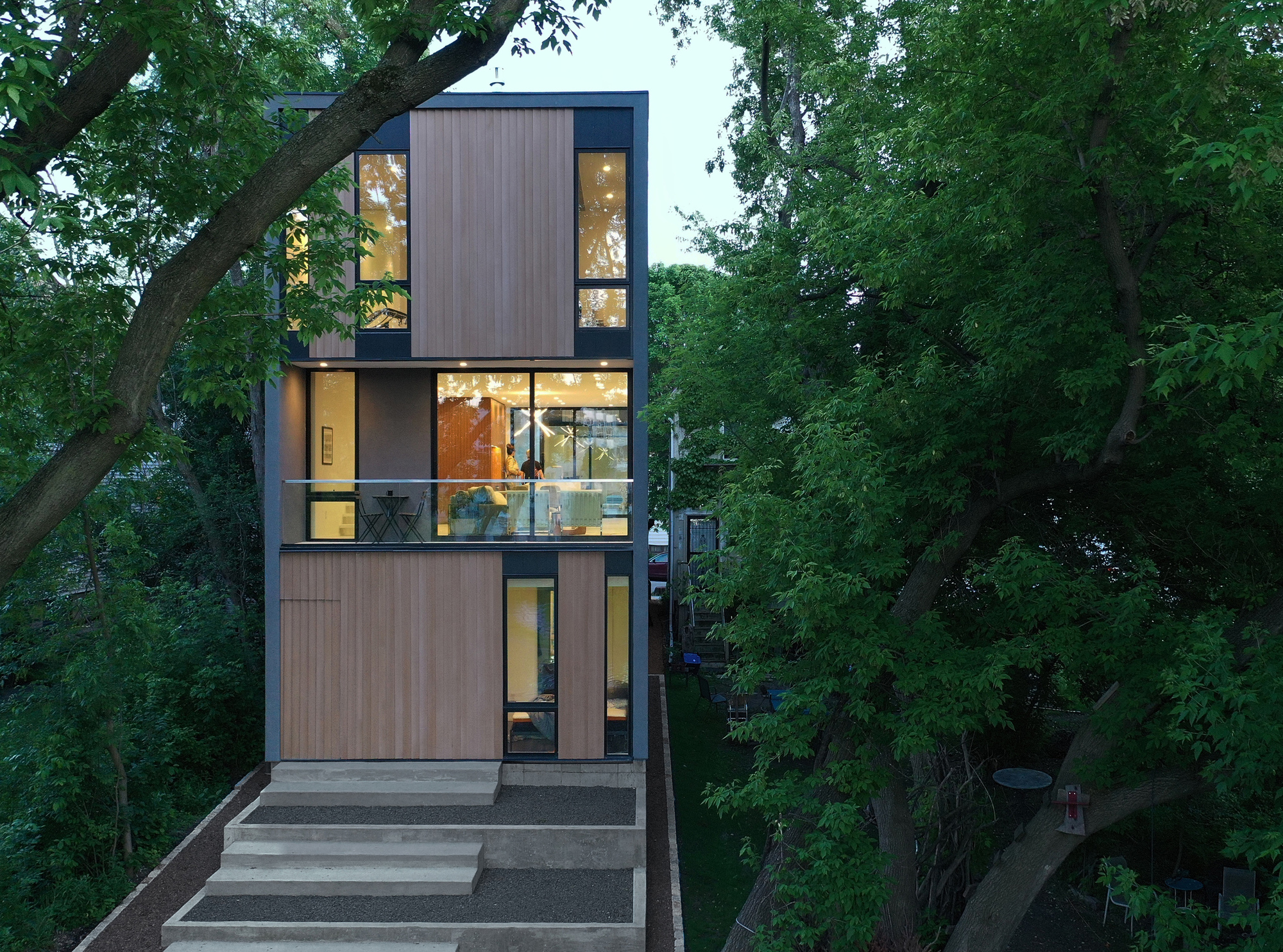 Narrow street facade with timber panels, lit glazing and a glass balcony rail above concrete steps, framed by green summer trees at dusk