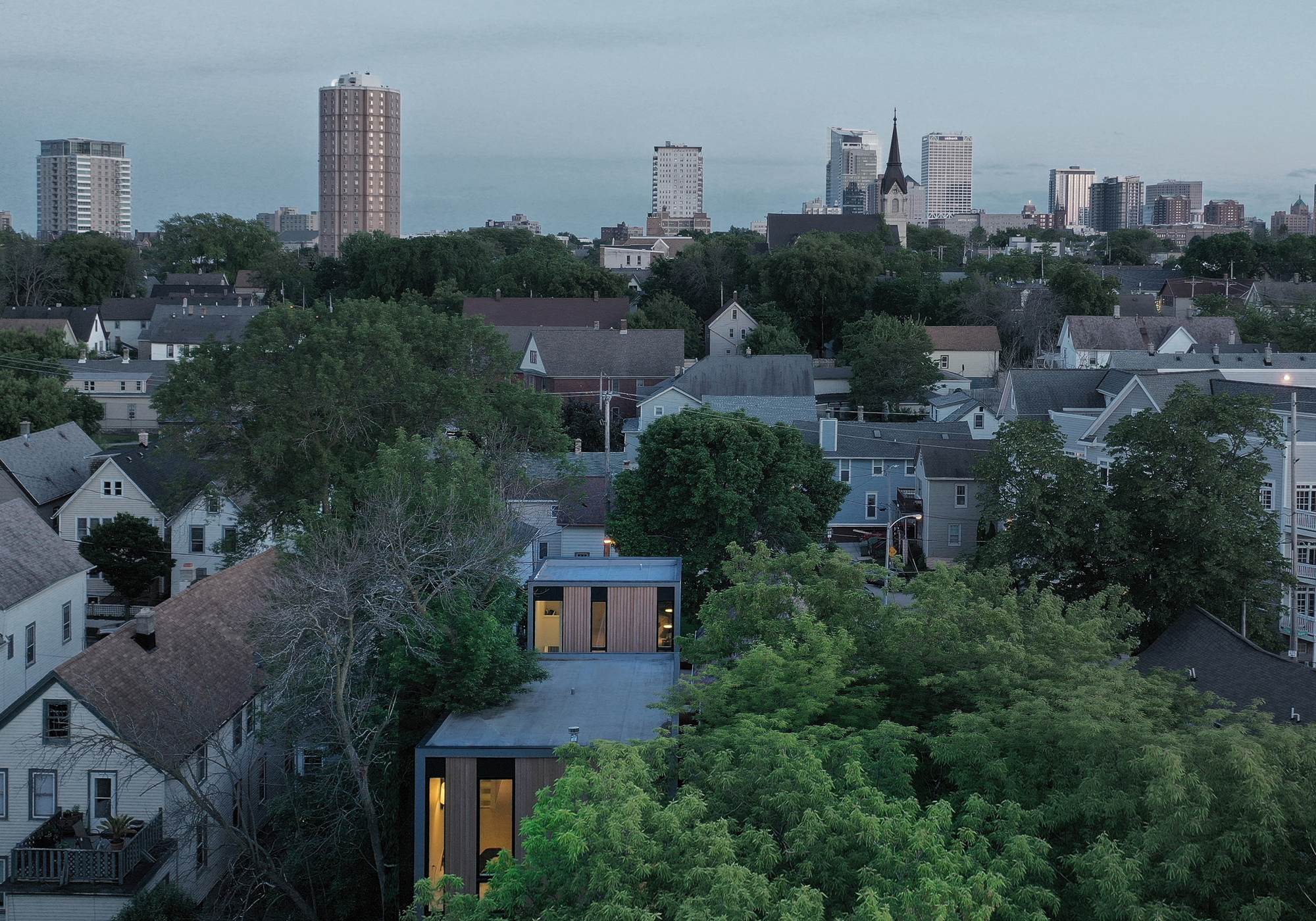 Aerial view of the small timber-clad house with glowing windows among leafy treetops and neighbouring gabled roofs, with the downtown skyline behind at dusk