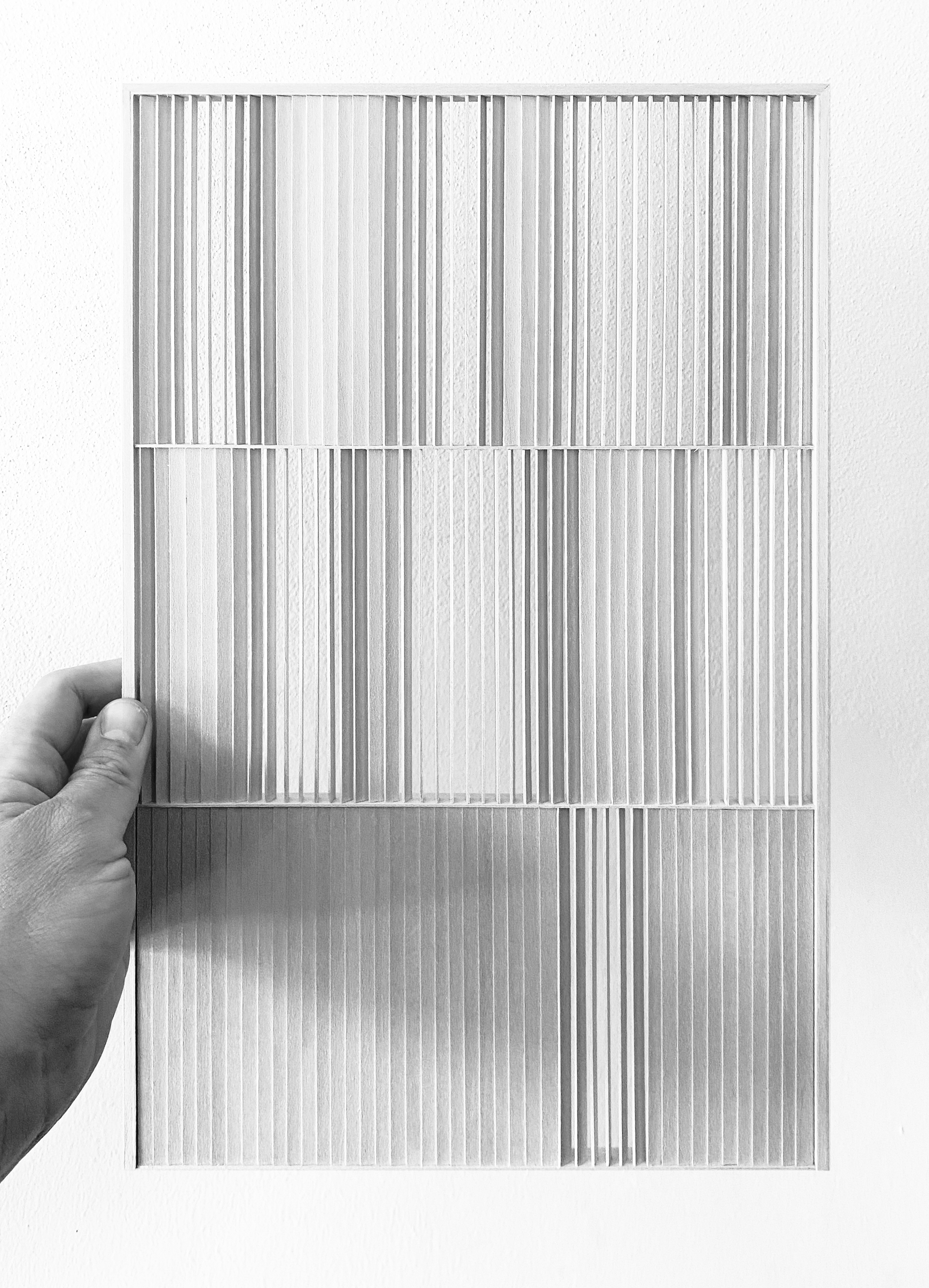 Hand holding a framed paper relief of vertical fins in three tiers, casting soft shadows against a white wall