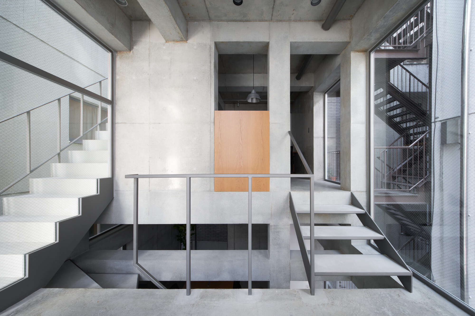Open concrete stairwell with grey steel handrails, a plywood panel and a glazed stair beyond, lit by soft daylight