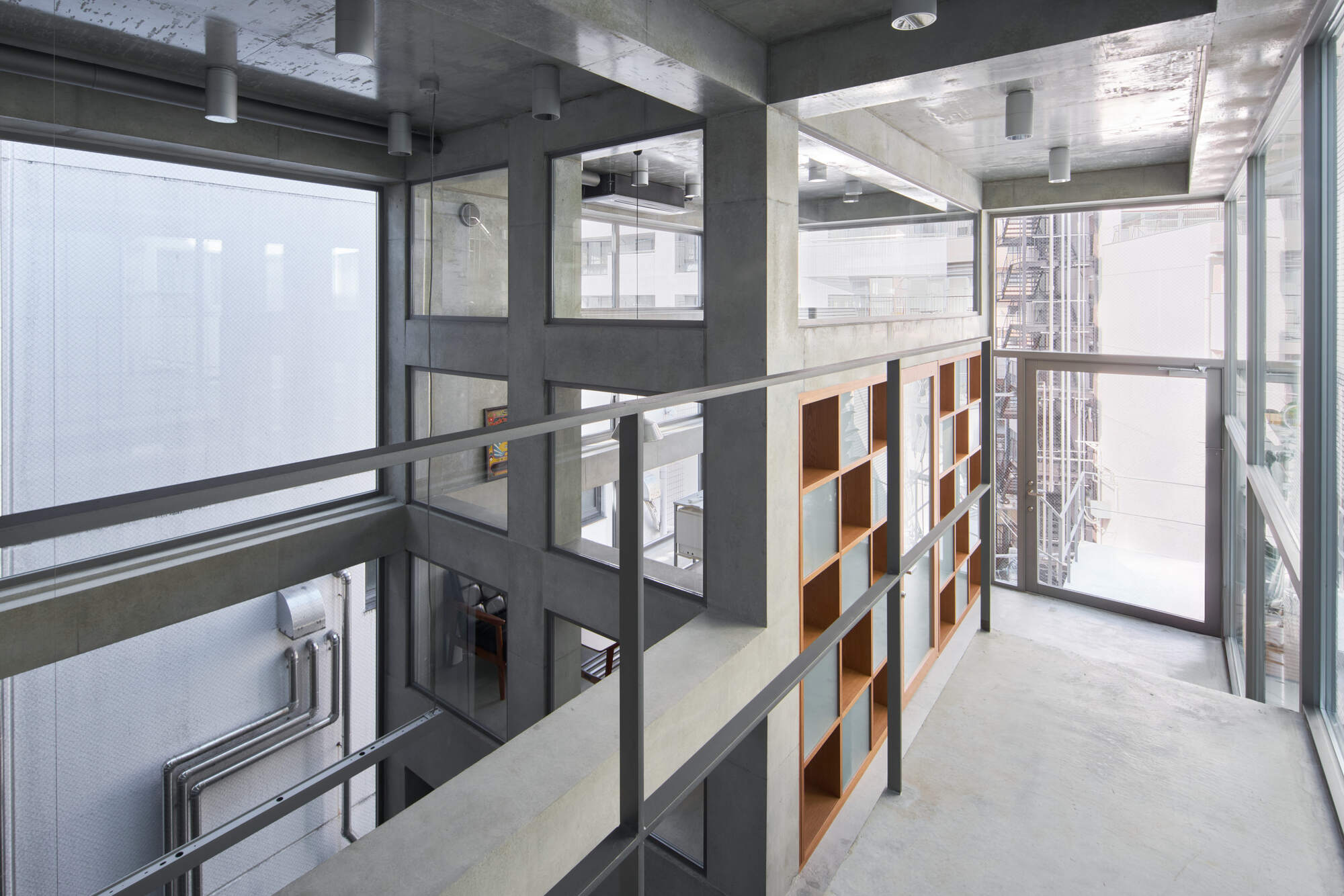 Concrete walkway with steel railing beside a built-in timber bookshelf, overlooking a gridded wall and neighboring fire escape in daylight