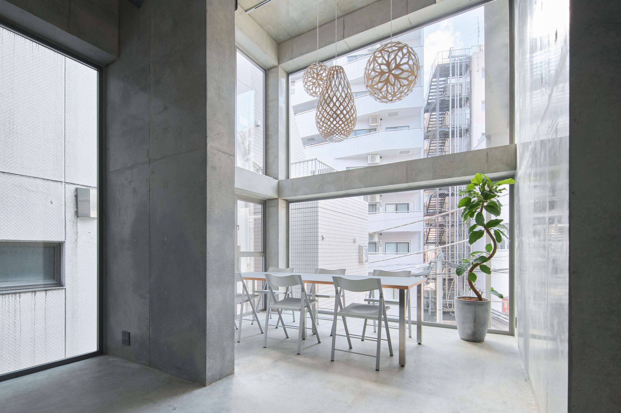Double-height room with folding chairs at a white table, a potted ficus and woven pendant lamps facing an adjacent apartment block