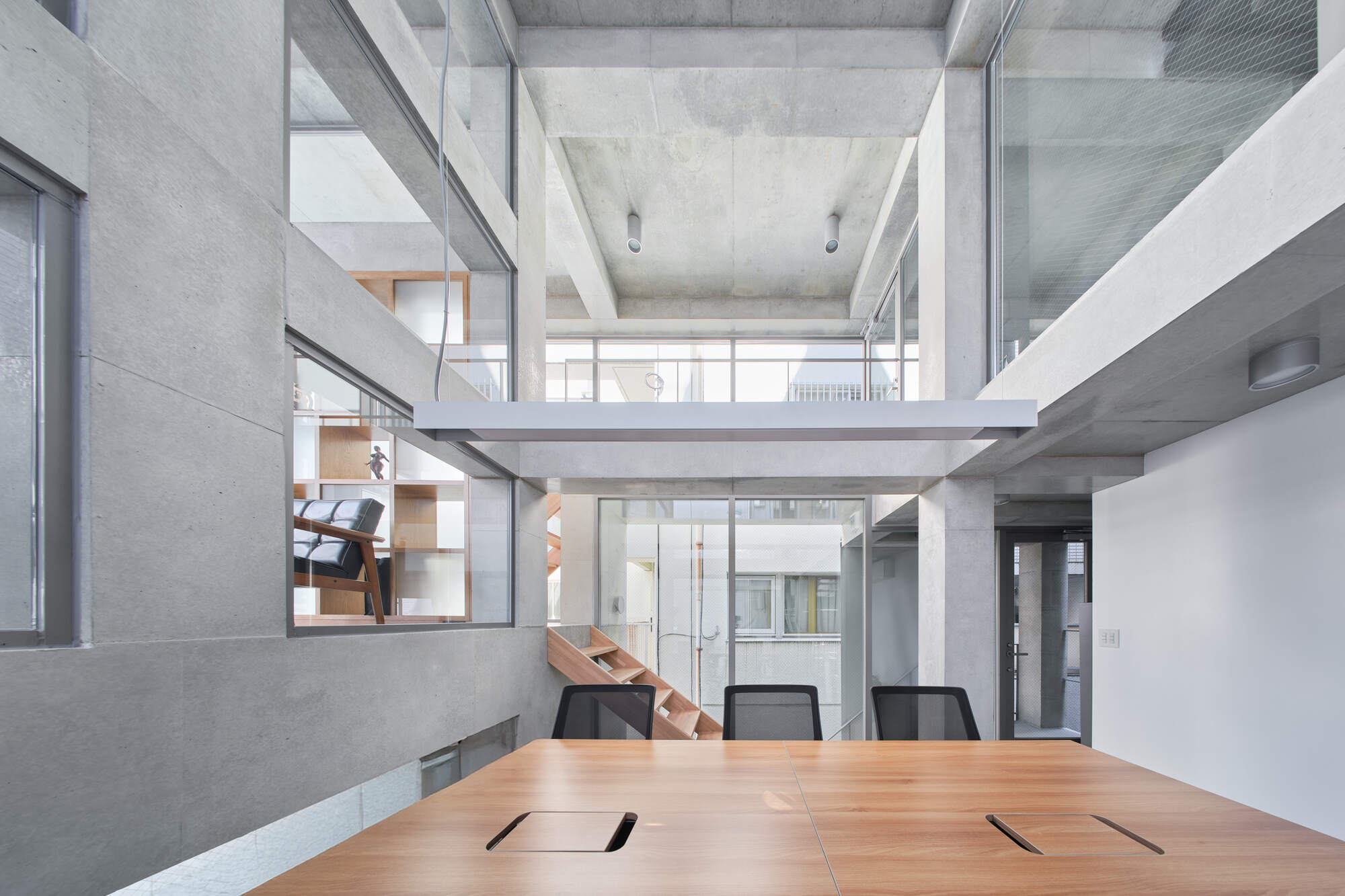 Wooden meeting table in a tall concrete atrium, with a timber stair and glazed lounge visible through openings in the grey wall