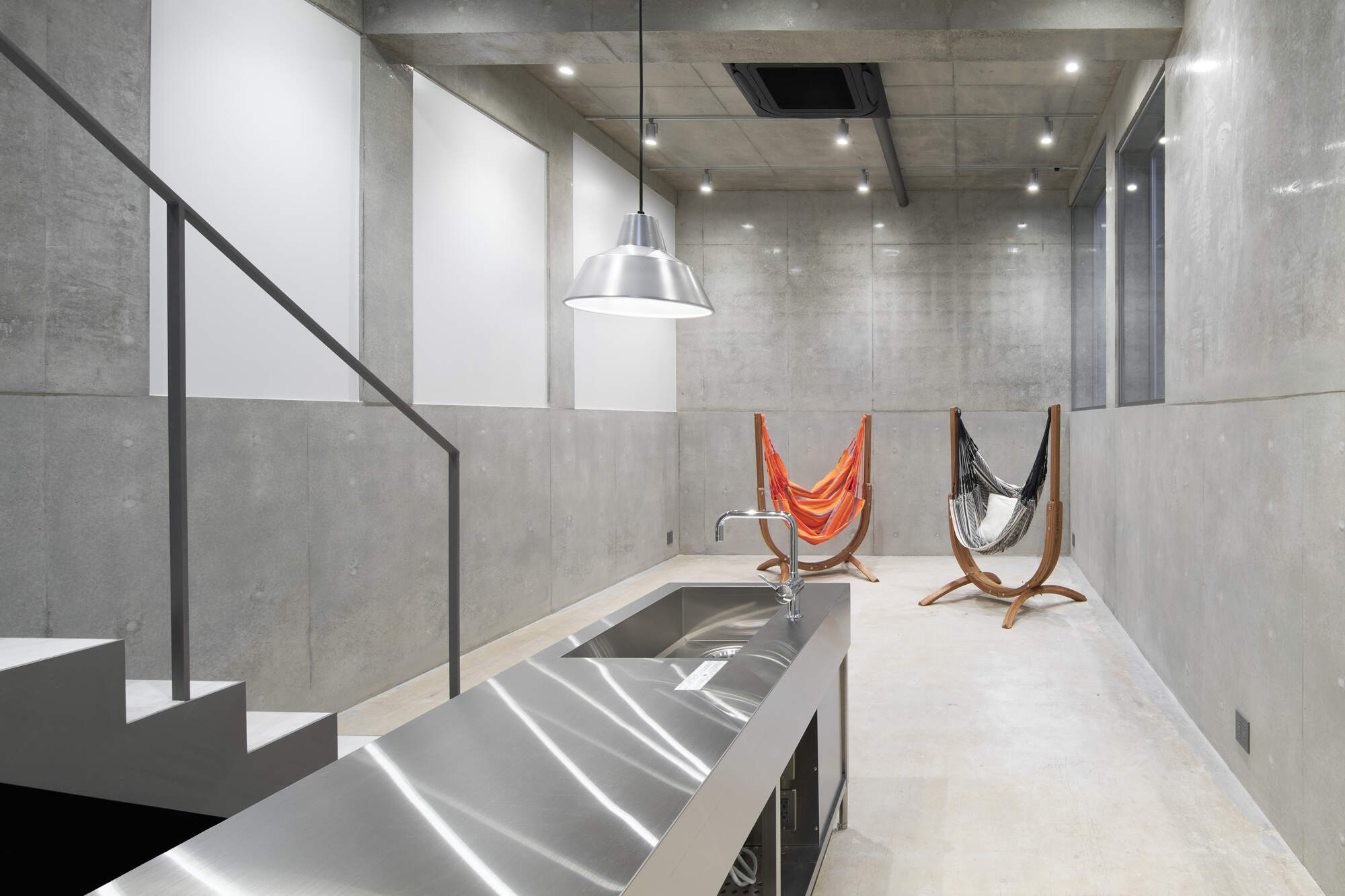 Stainless steel kitchen counter in a concrete basement room with two hammock chairs, a pendant lamp and a steel-railed stair