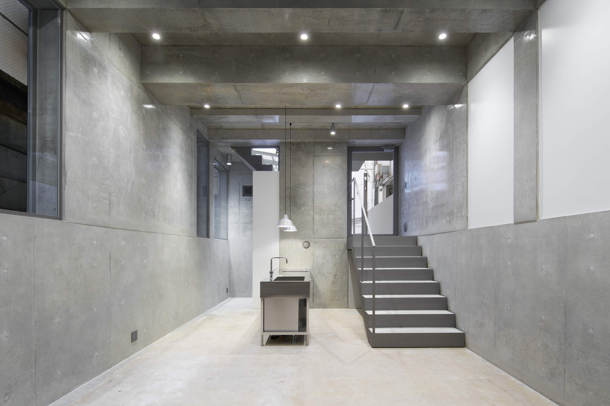 Concrete-walled ground floor room with a steel kitchen unit and a gray staircase rising toward a glazed door, lit by ceiling spotlights