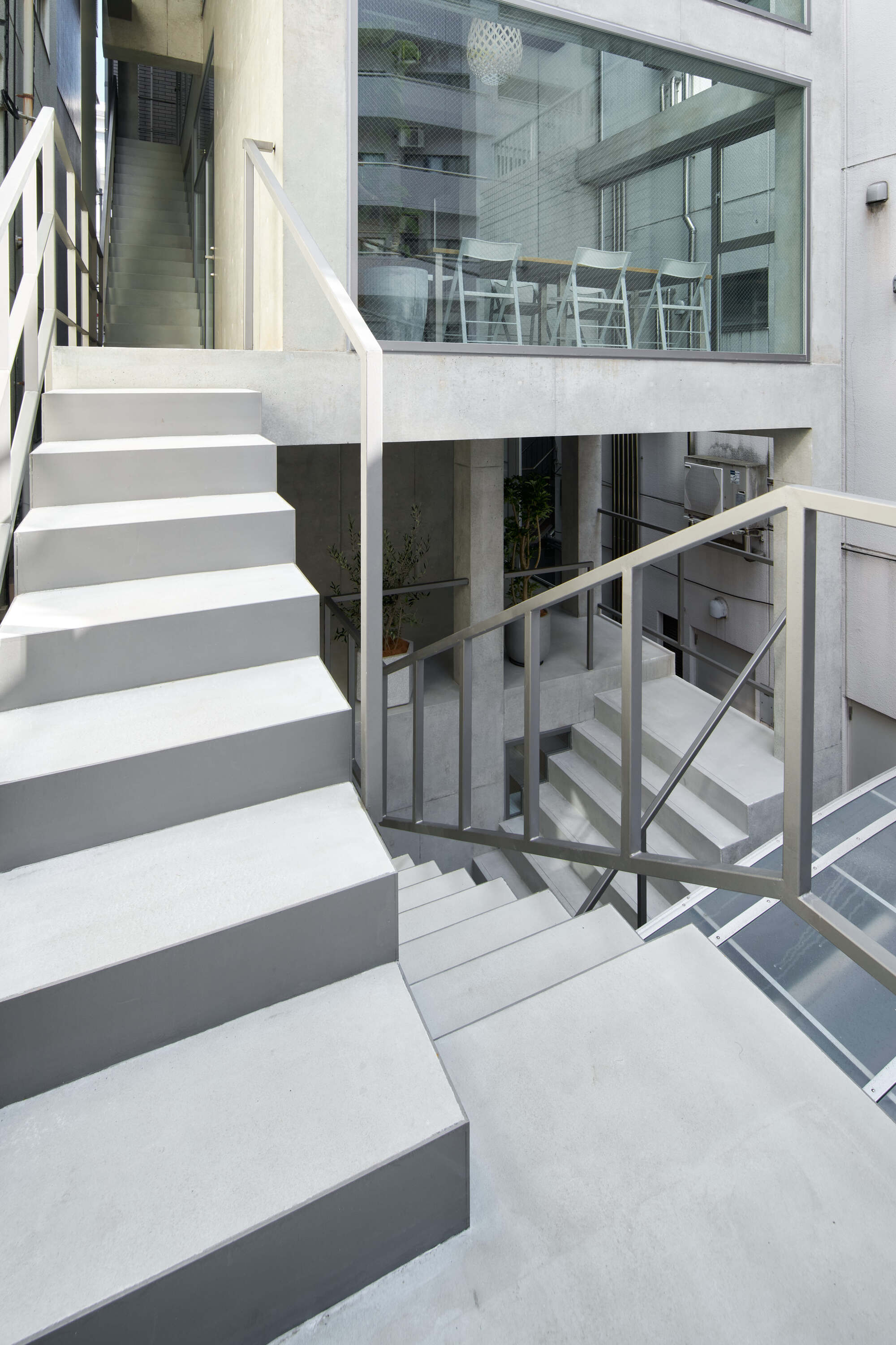 Exterior concrete stair flights with thin steel railings stepping down past a glazed meeting room and potted plants in soft daylight