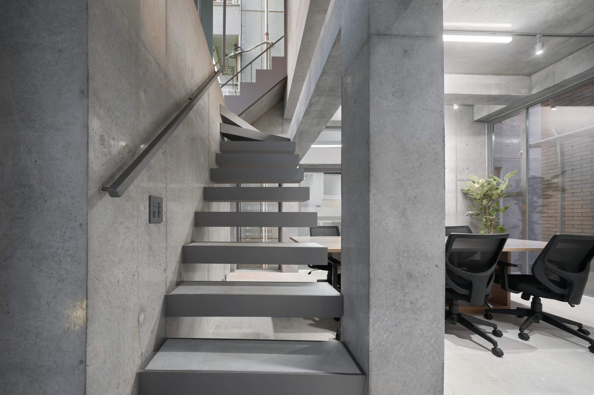 Cantilevered steel-tread stair beside a concrete column, with a meeting table and mesh-back chairs visible through the opening