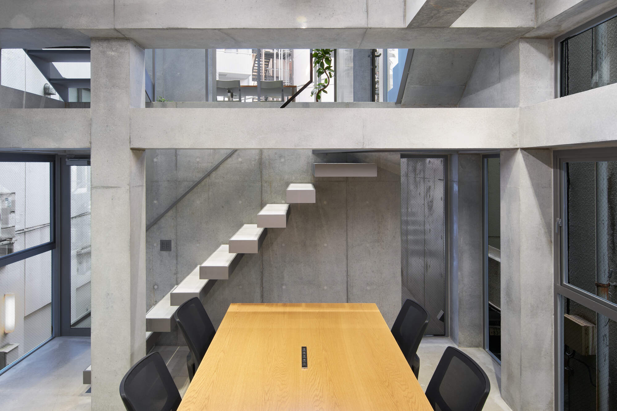 Wooden meeting table below a floating concrete stair, framed by concrete columns and beams with a plant on the upper level