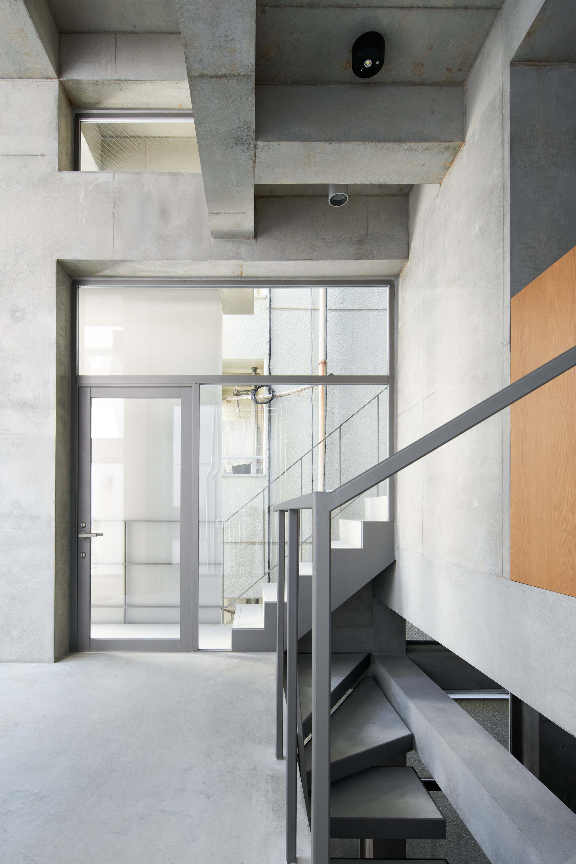 Concrete stair landing with a gray-framed glass door, exposed ceiling beams and a steel balustrade over a spiral of dark treads