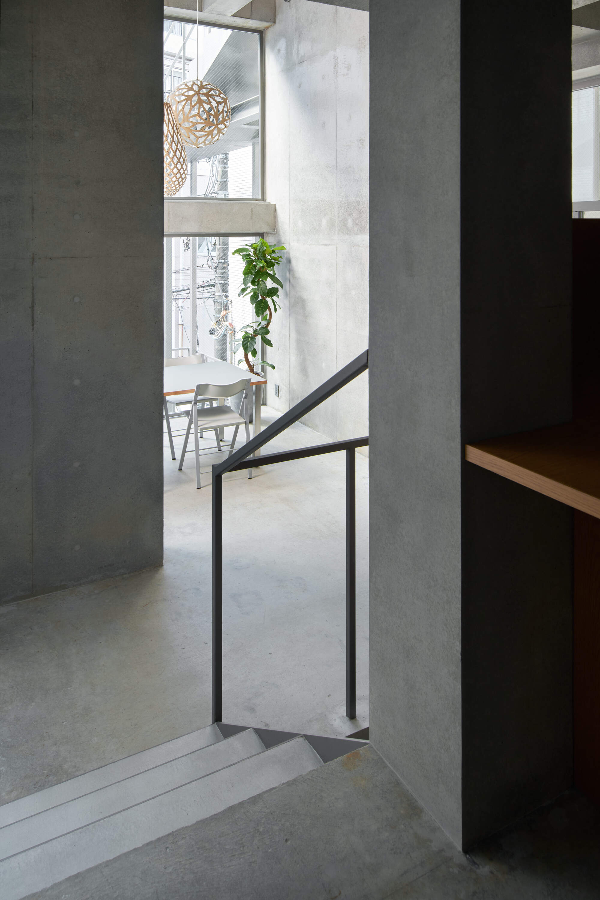 Concrete stair head with a black steel handrail looking toward a white table, chairs, a potted plant and a woven pendant lamp