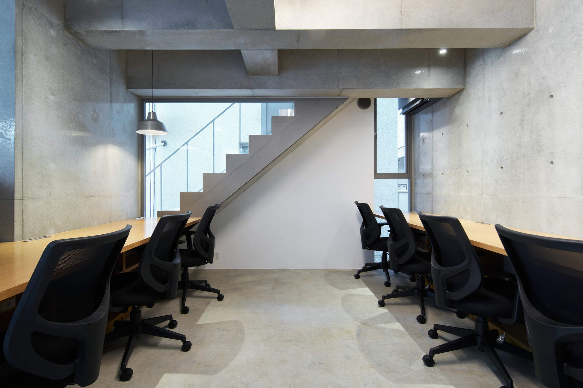 Office room with rows of black mesh chairs at timber desks, facing an open stair behind a window