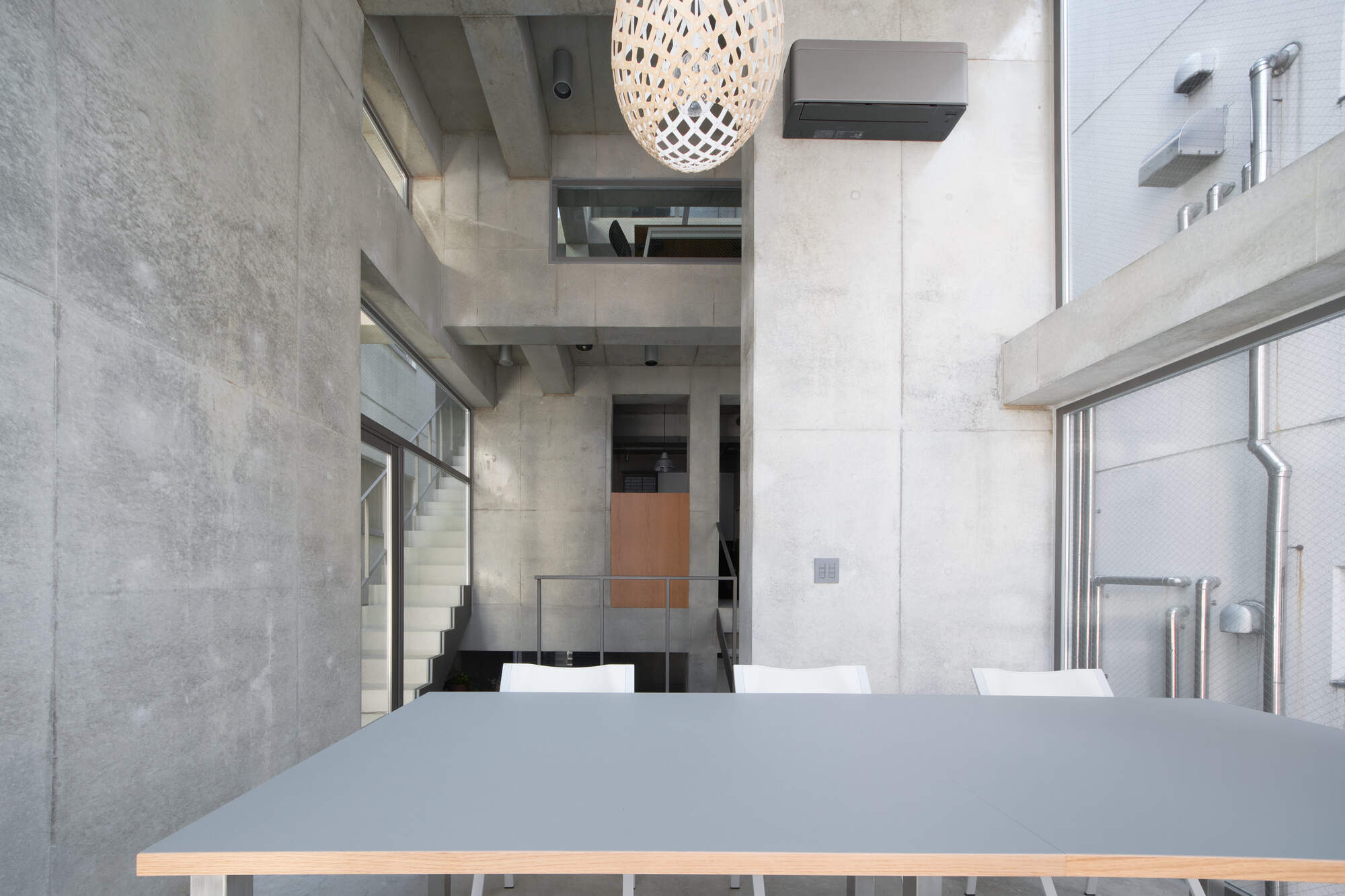 Grey laminate table with white chairs in a concrete atrium, a woven pendant lamp hanging overhead in daylight