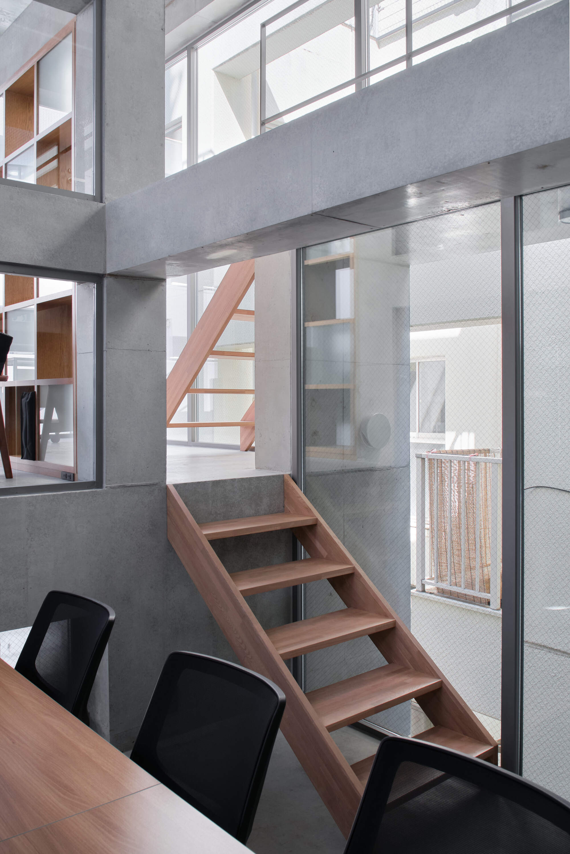 Open timber stair rising beside a glass wall, with office chairs and a shelving unit in the foreground
