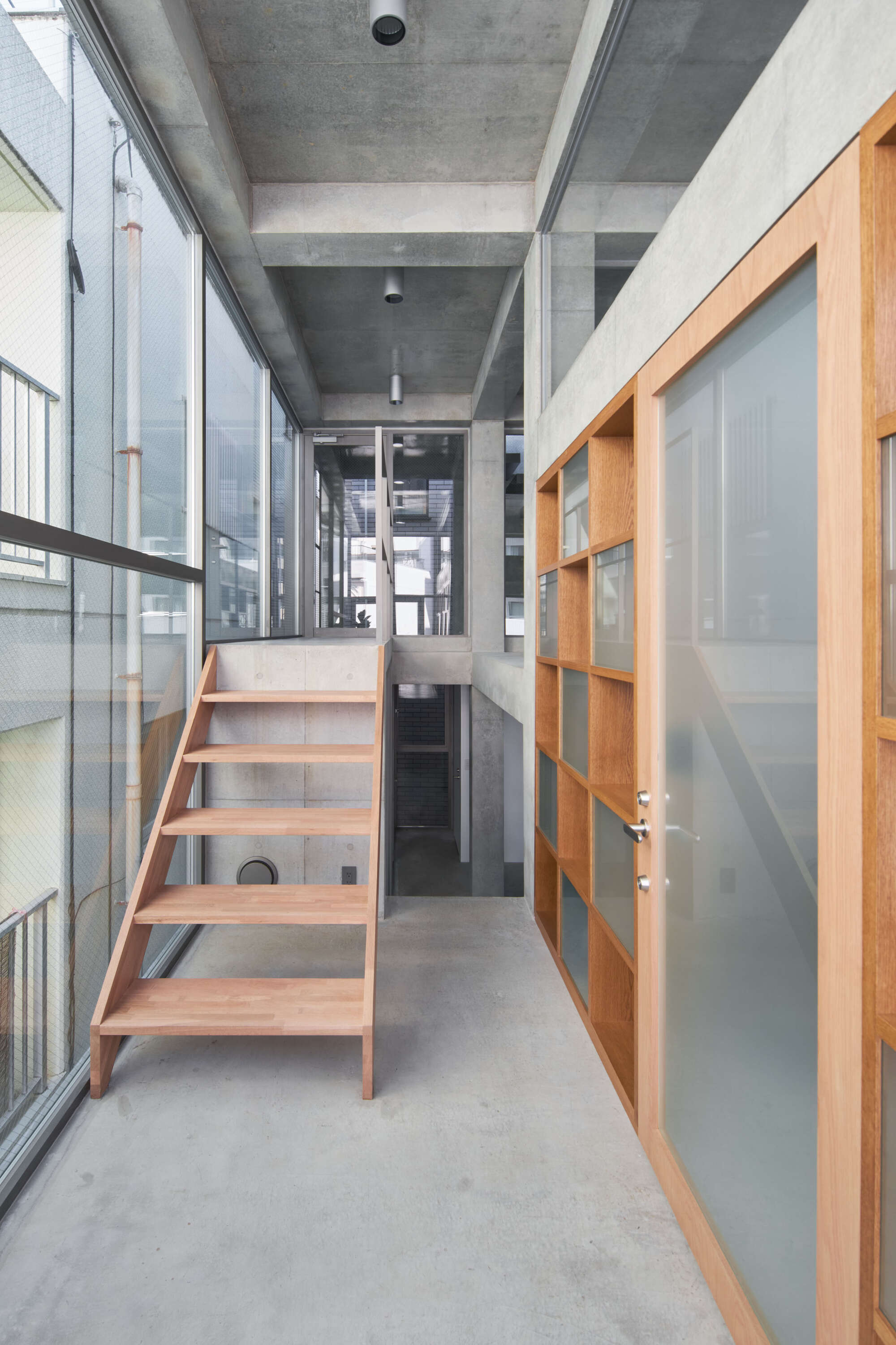 Narrow concrete corridor with a timber stair, glass wall on one side and oak shelving with frosted door on the other