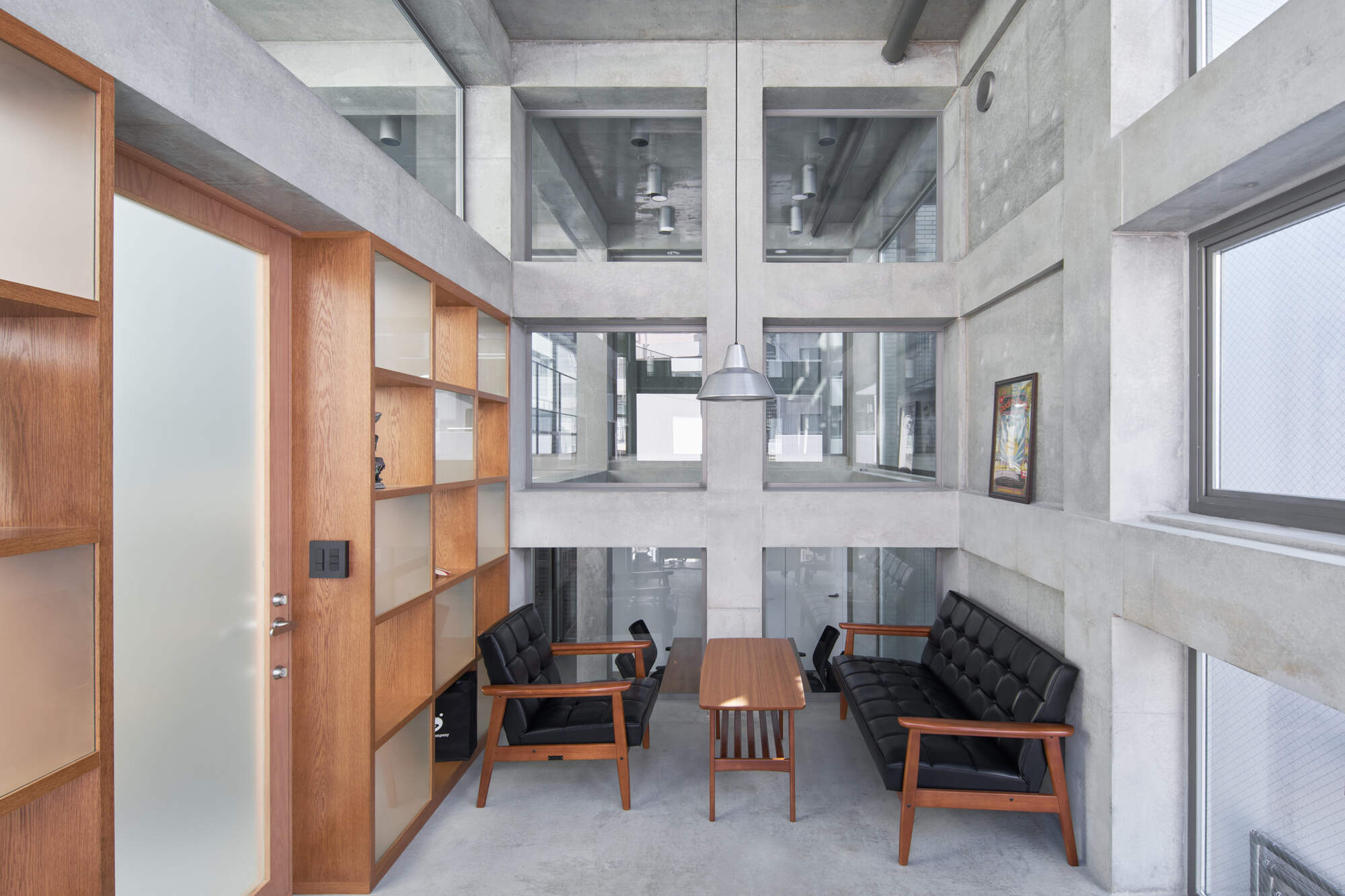 Double-height lounge with black leather sofa, wooden side table and oak shelving against a gridded concrete wall