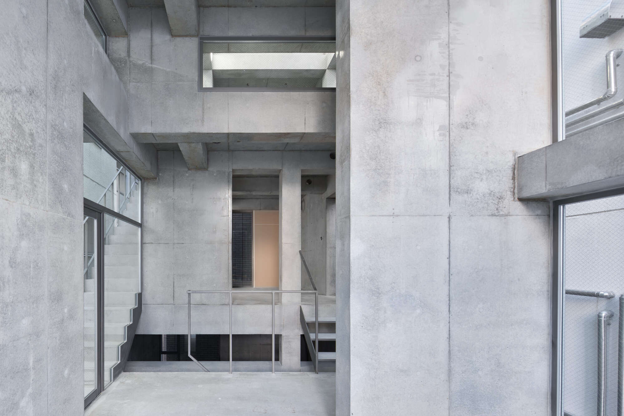Bare concrete atrium with a steel railing at a landing, a glazed stair at left and pale daylight above