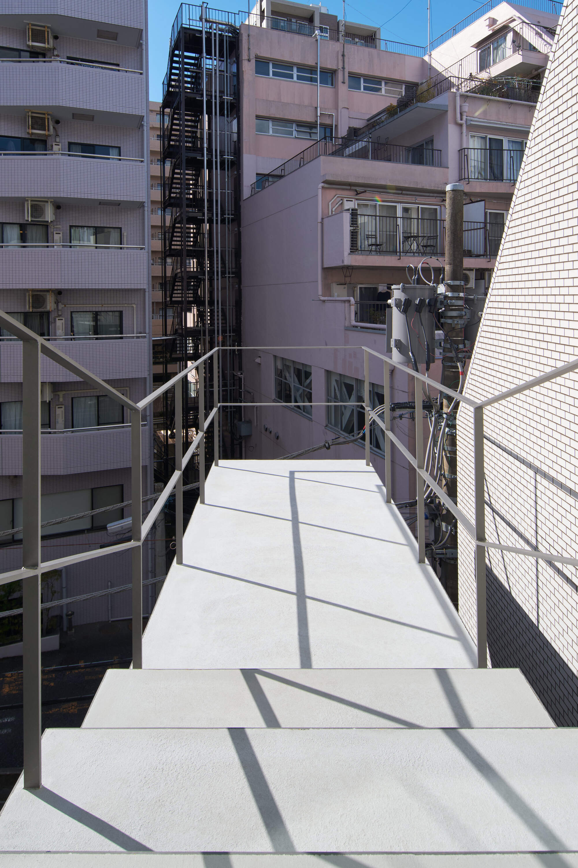 Open exterior steel stair landing with thin metal railings, casting shadows in sunlight toward neighboring apartment blocks and a fire stair