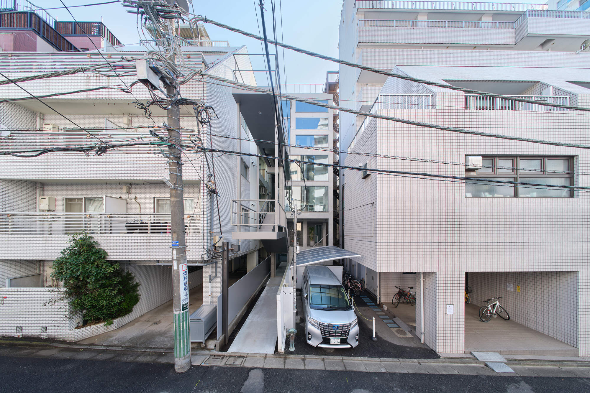 Street view of a narrow tiled building between neighbors, with a parked van, bicycles and utility wires under a pale sky