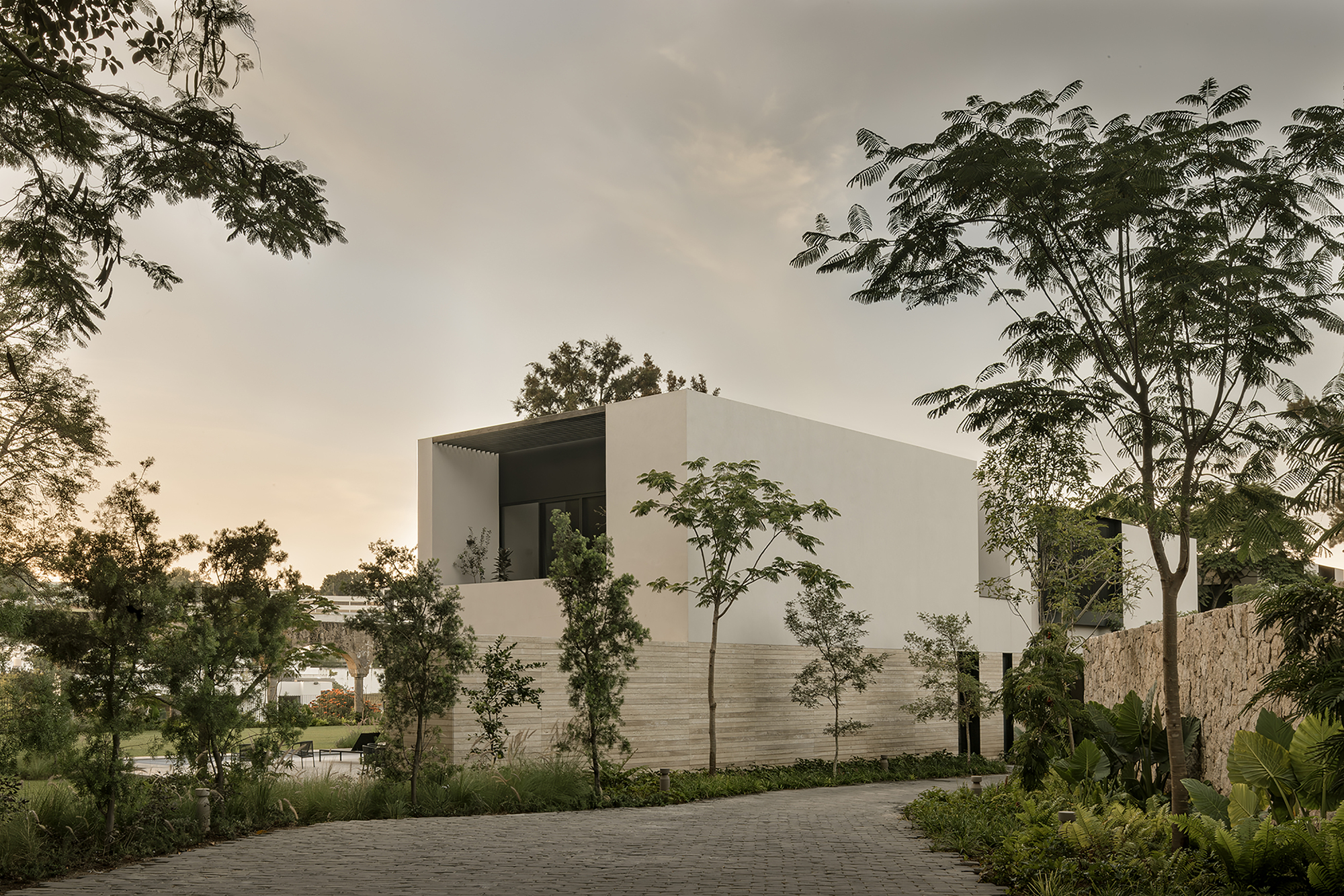 White box volume with a recessed upper terrace and banded stone base, seen from a paved drive at dusk