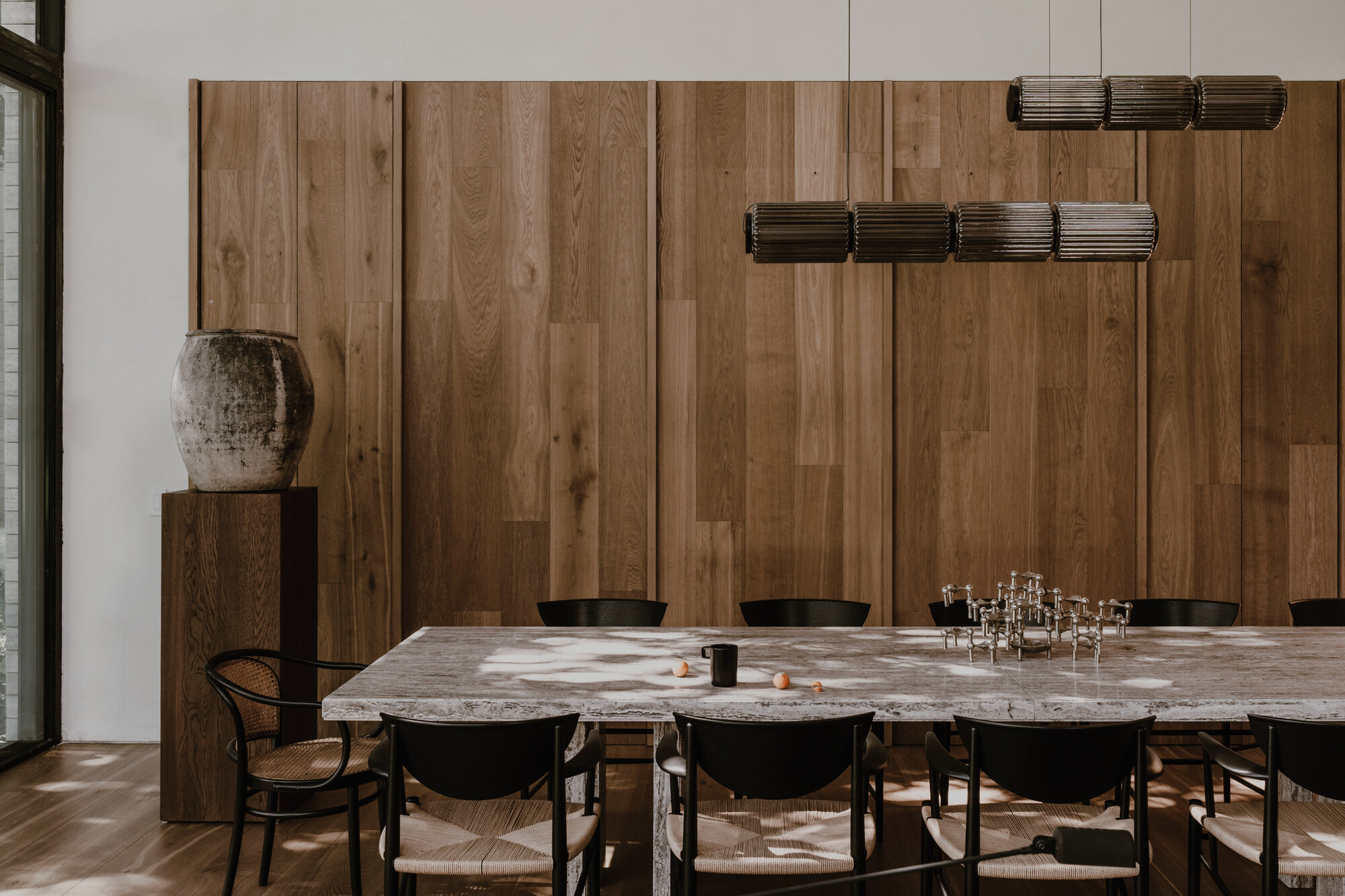Long stone dining table with black rattan chairs against an oak panel wall, with sunlight patches on the tabletop