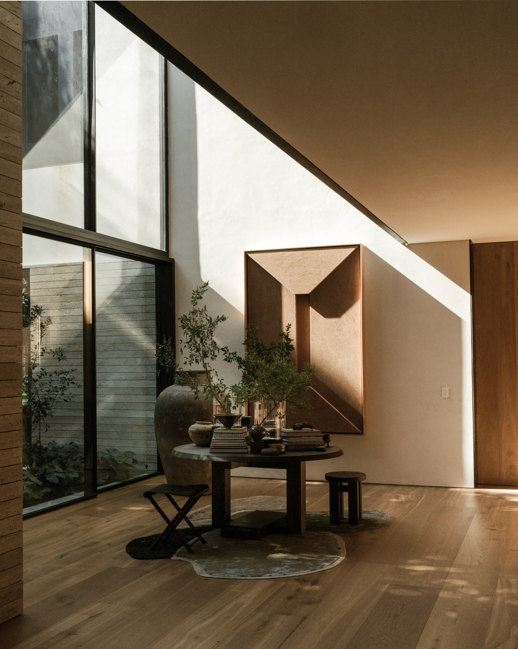 Entry hall with a round table and stools beneath a sloped ceiling, with late sun cutting across a plaster wall