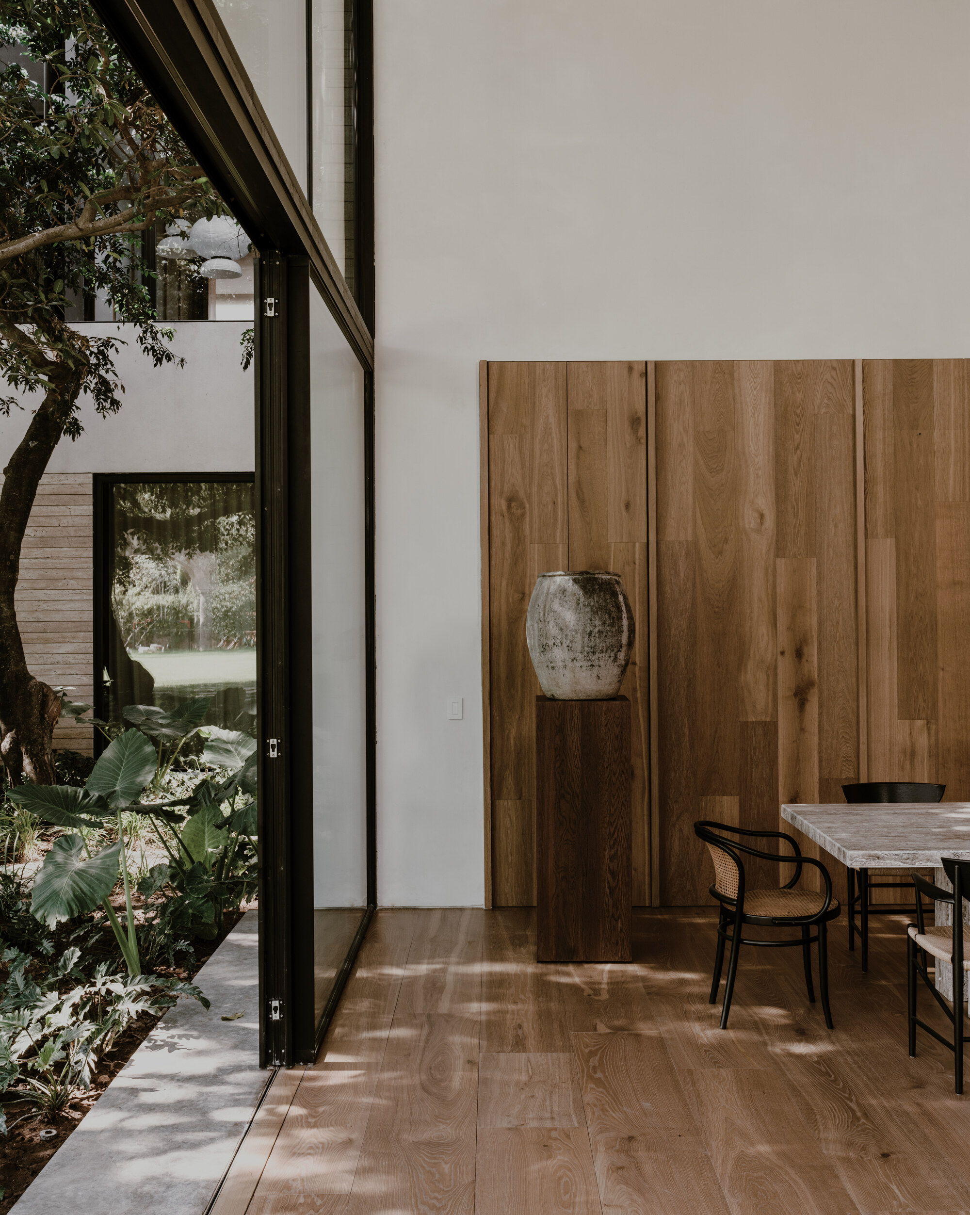 Oak wall panels and a stone urn on a wooden plinth beside a glazed corner opening onto a planted courtyard, with dappled sunlight on the floor