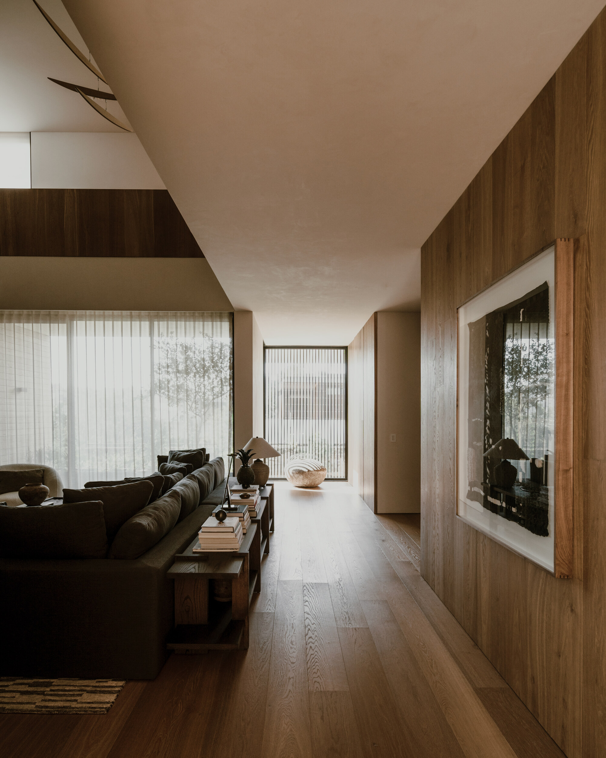 Long oak-clad hallway running past a linen sofa and sheer-curtained windows, with soft daylight falling across the wide plank floor