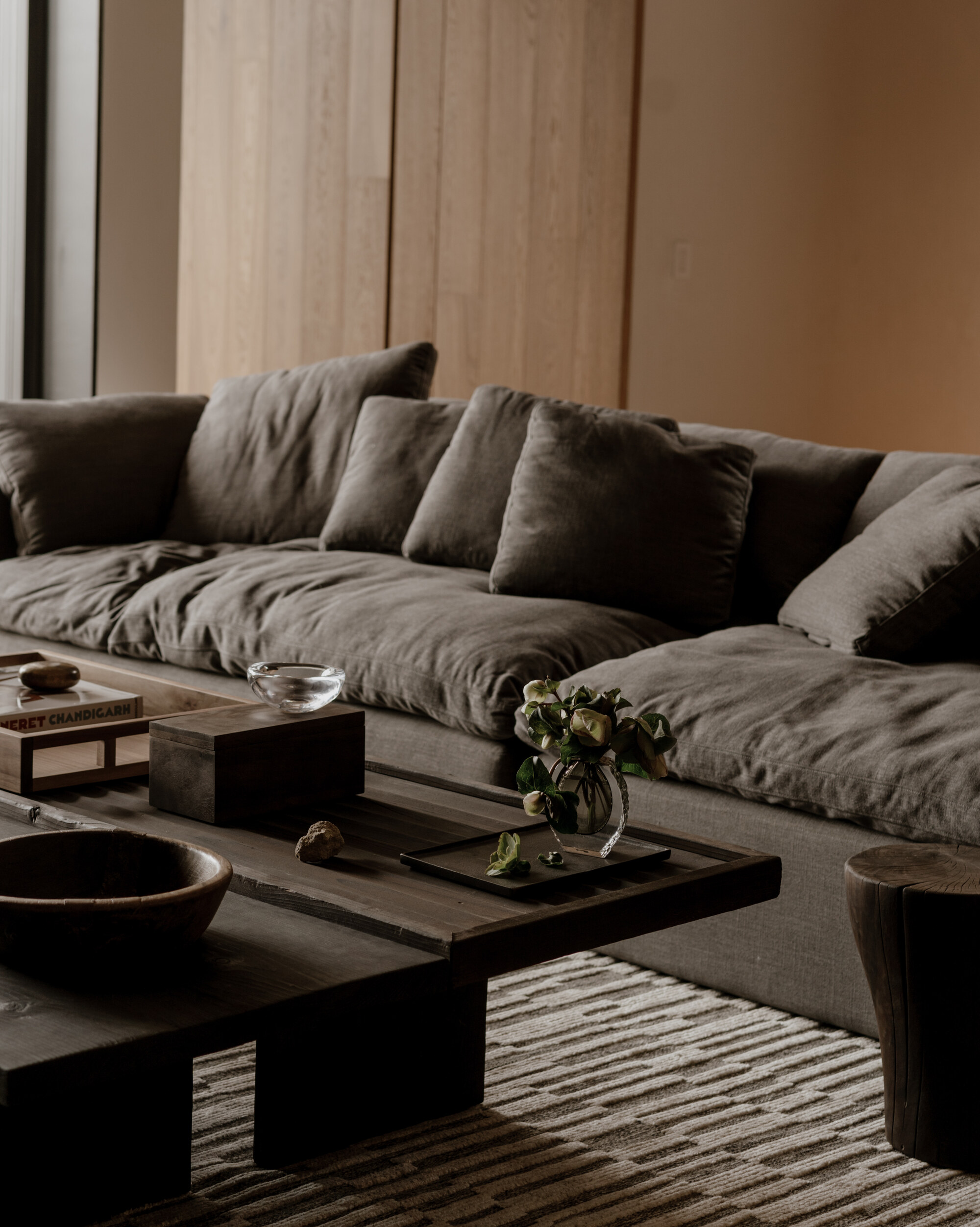 Close-up of a deep grey linen sofa with loose cushions behind a low dark wood coffee table holding a bowl, tray and flowers