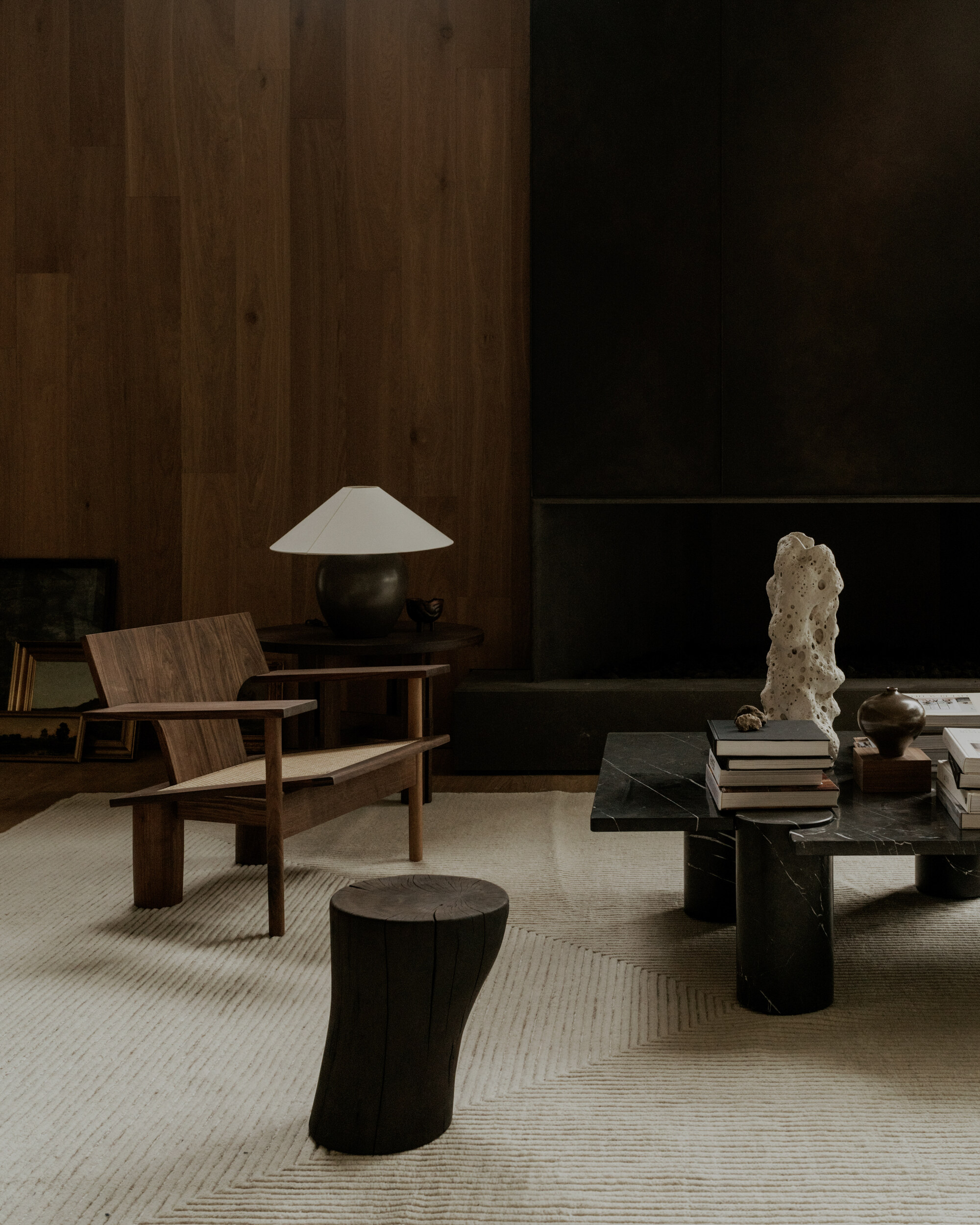 Walnut lounge chair, black stump stool and marble-legged coffee table on a cream rug before a dark timber wall in low light