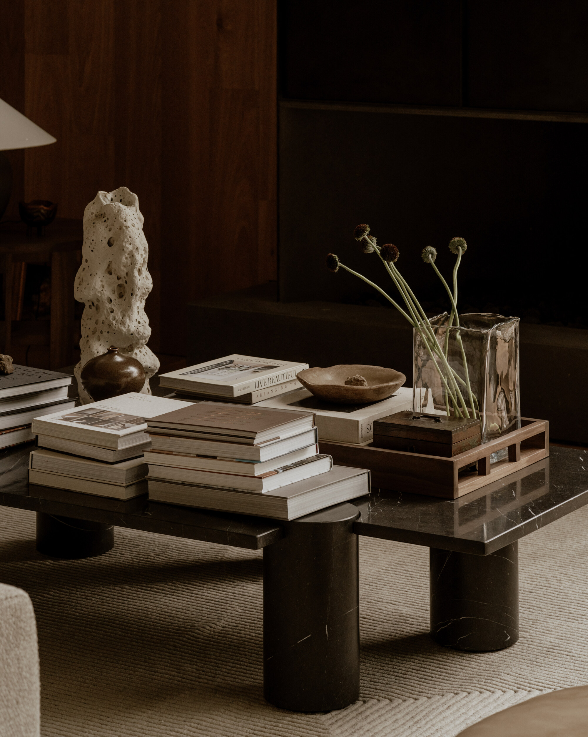 Coffee table with stacked books, a porous white sculpture, a glass vase of dried stems and a wooden tray in dim light