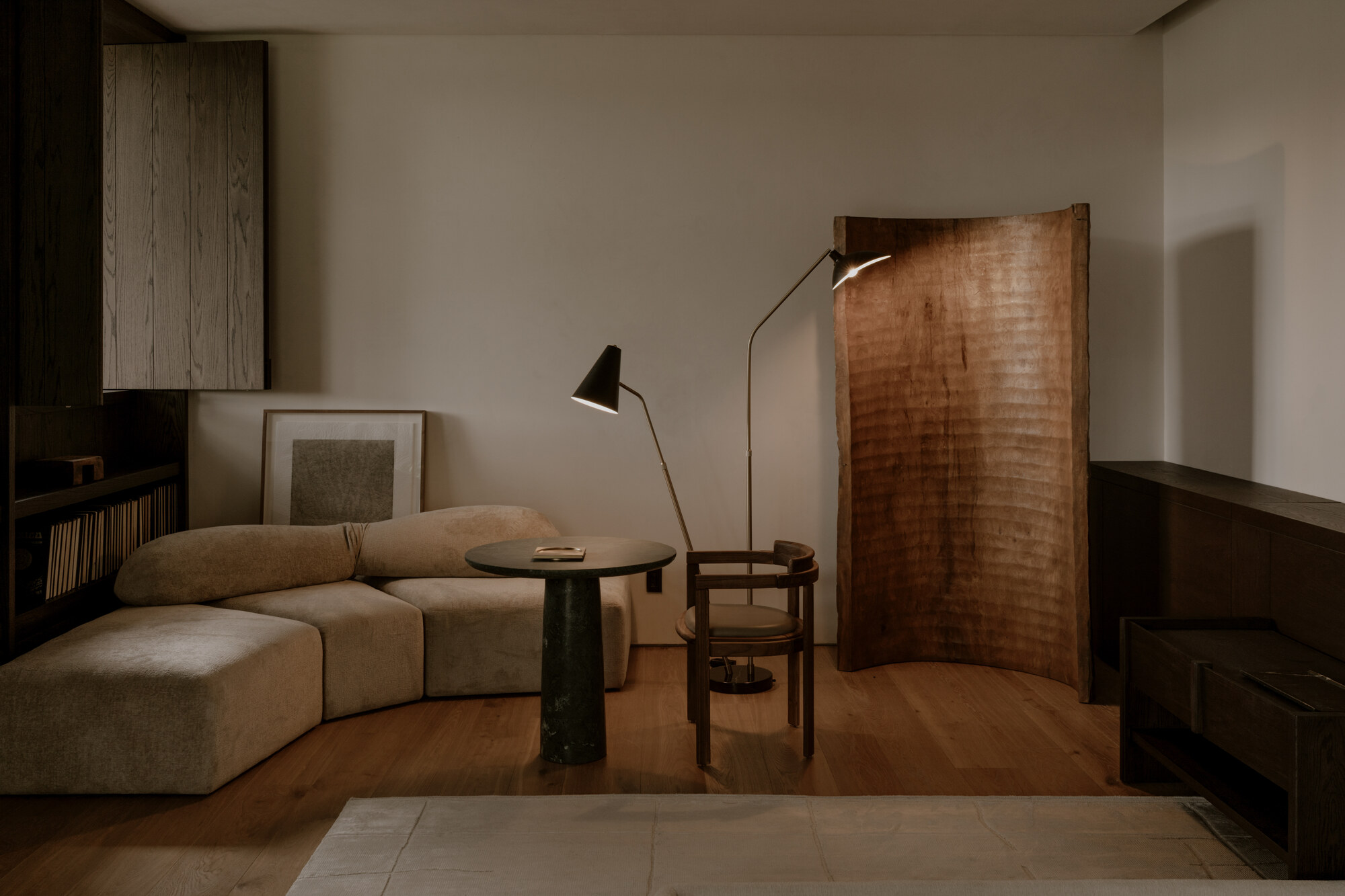 Curved sofa, round stone-based table and carved wooden screen lit by two black floor lamps against a pale plaster wall at dusk
