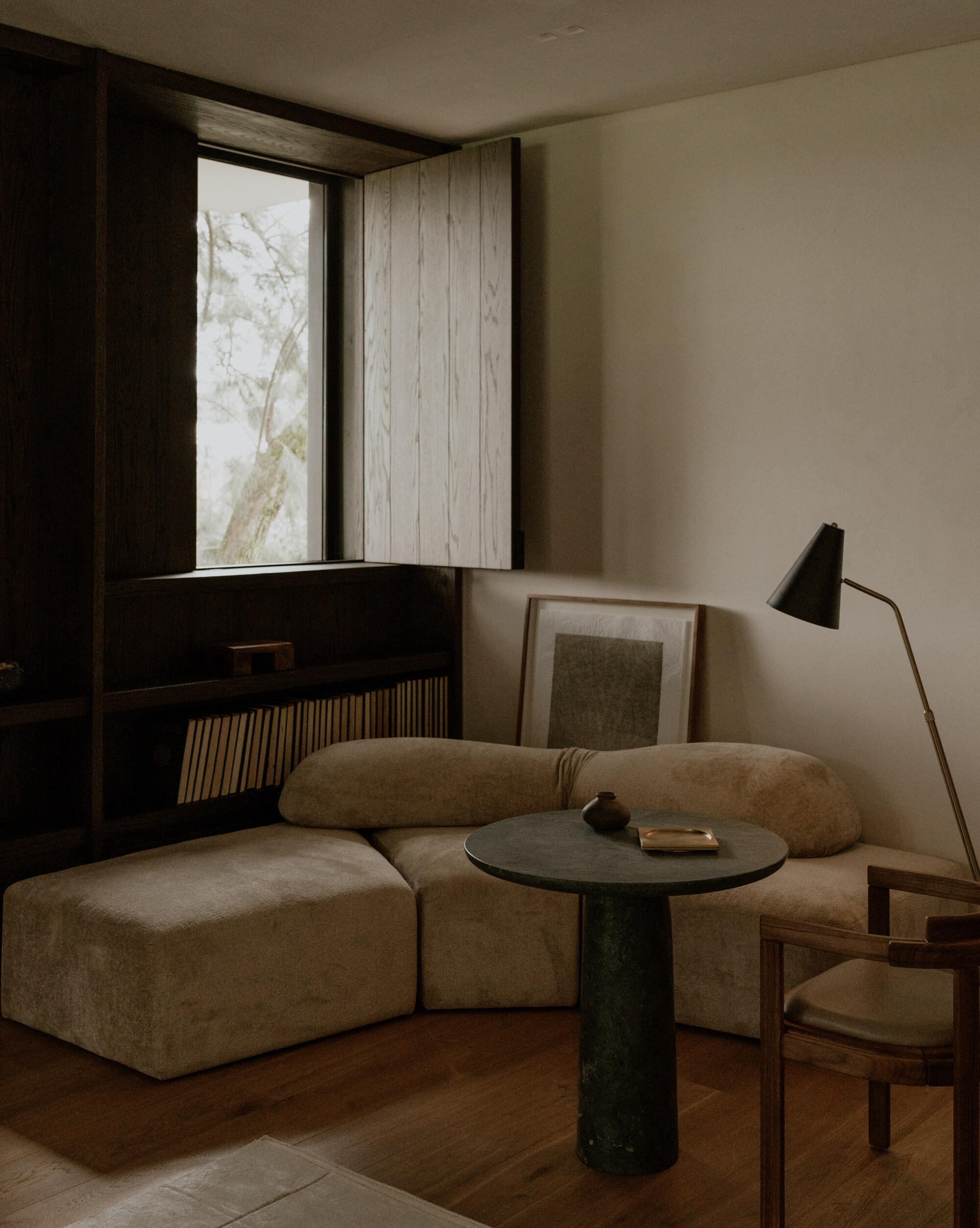 Curved beige sofa and round stone-topped table beside a dark timber bookshelf and open wooden shutter, with tree branches glowing in the window