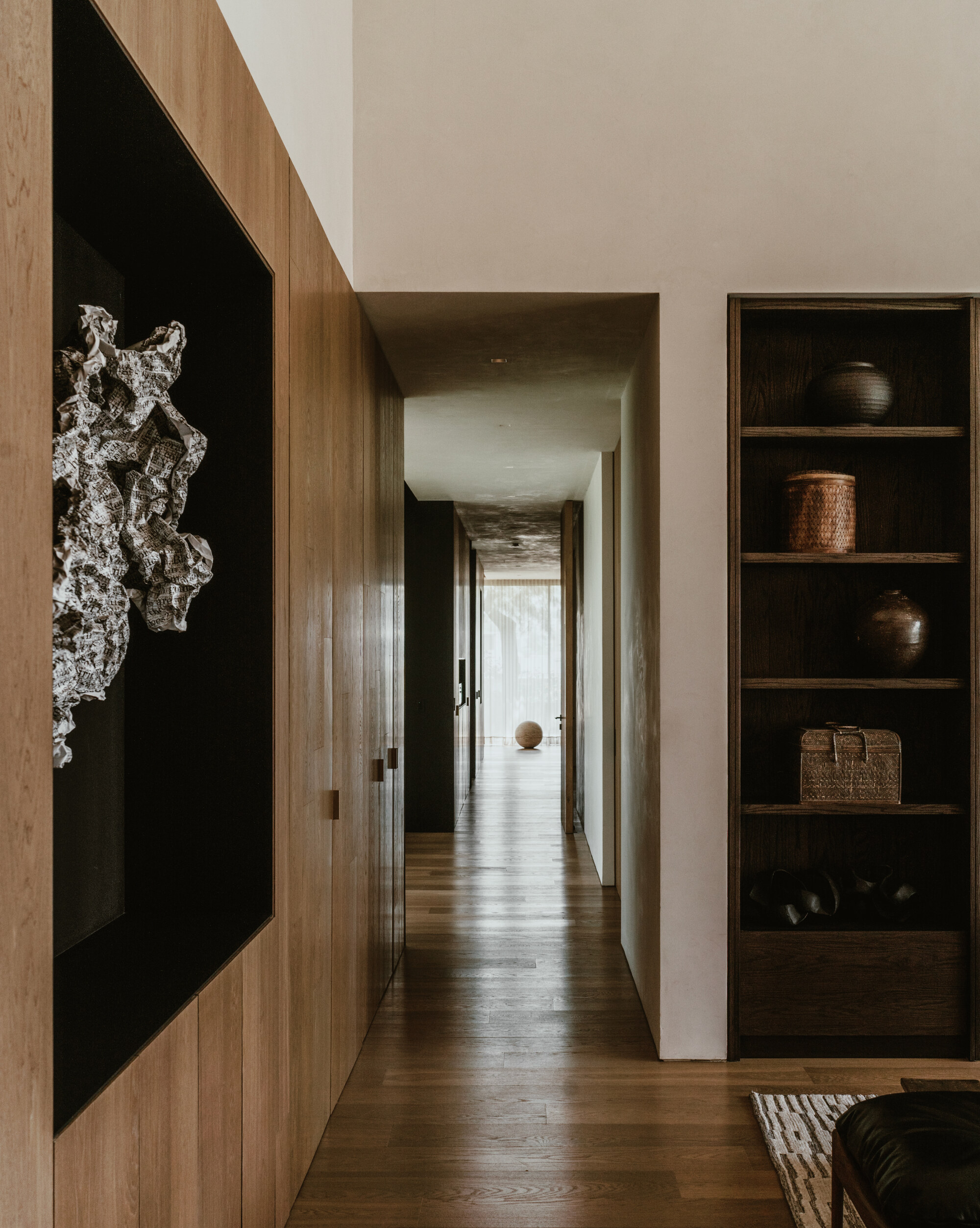 Long oak-floored corridor lined with a recessed sculpture niche and a dark shelf of vessels, ending in a bright curtained window