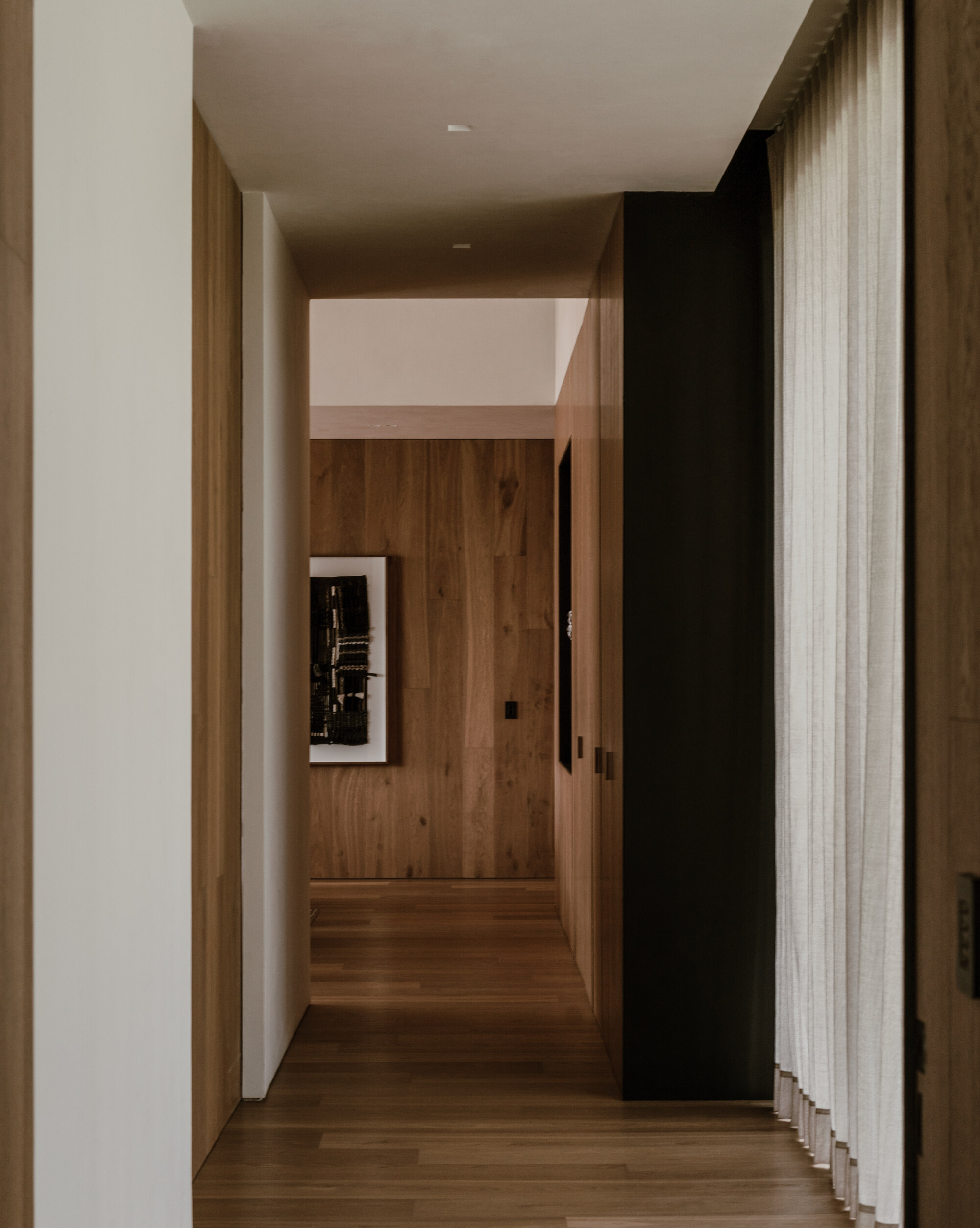 Narrow hallway of oak panelling and white plaster walls, with a framed artwork at the far end and a sheer curtain at right