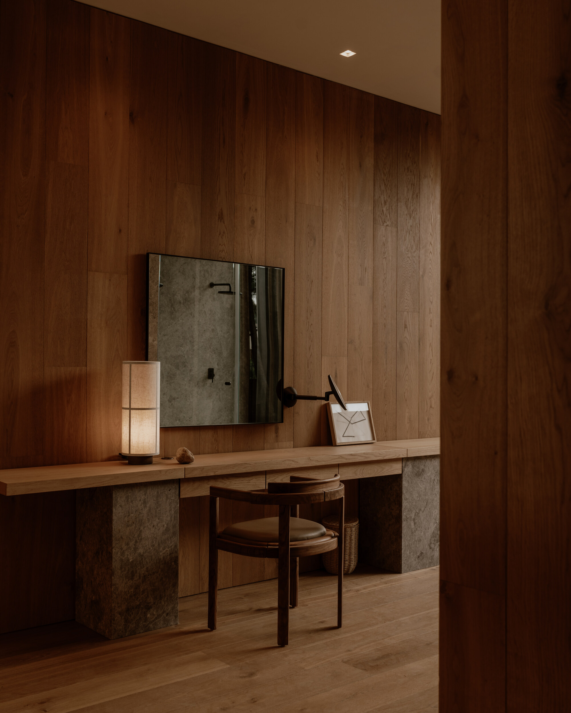 Timber dressing counter on two stone piers, with a wall mirror, paper lantern and round-backed wooden chair in warm lamplight