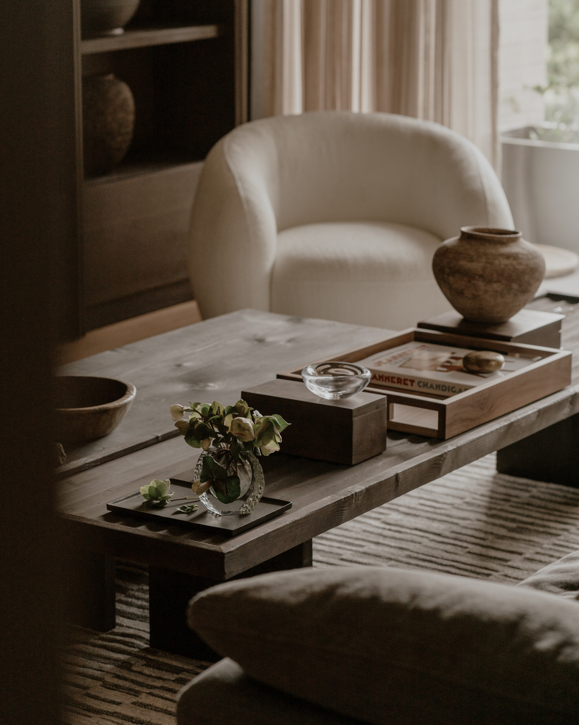 Low wooden coffee table with a tray, clay vase and small flower arrangement, in front of a white boucle armchair by a curtained window