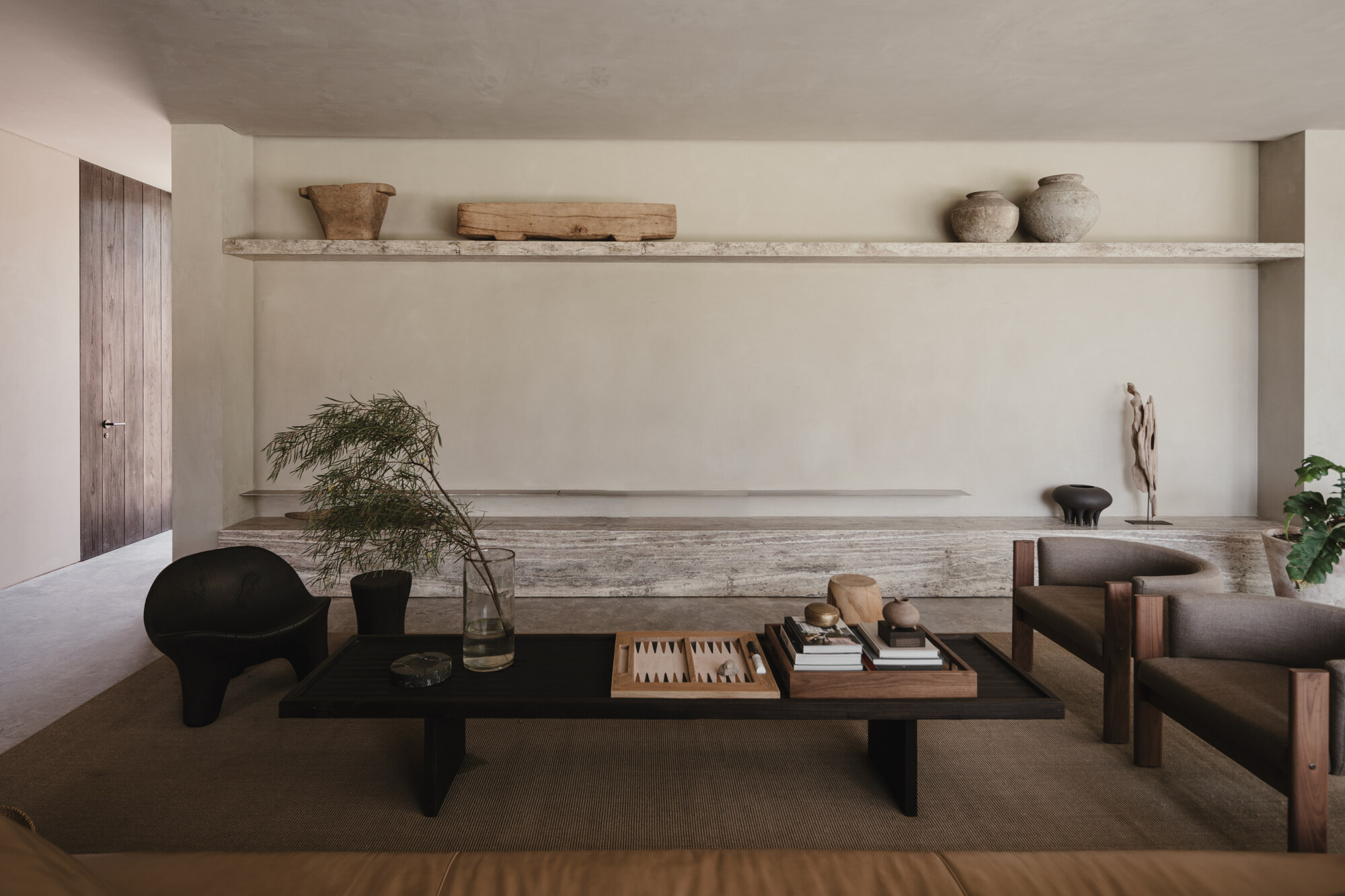 Living area with a travertine shelf holding clay vessels above a low dark coffee table, backgammon board and leather sofa edge
