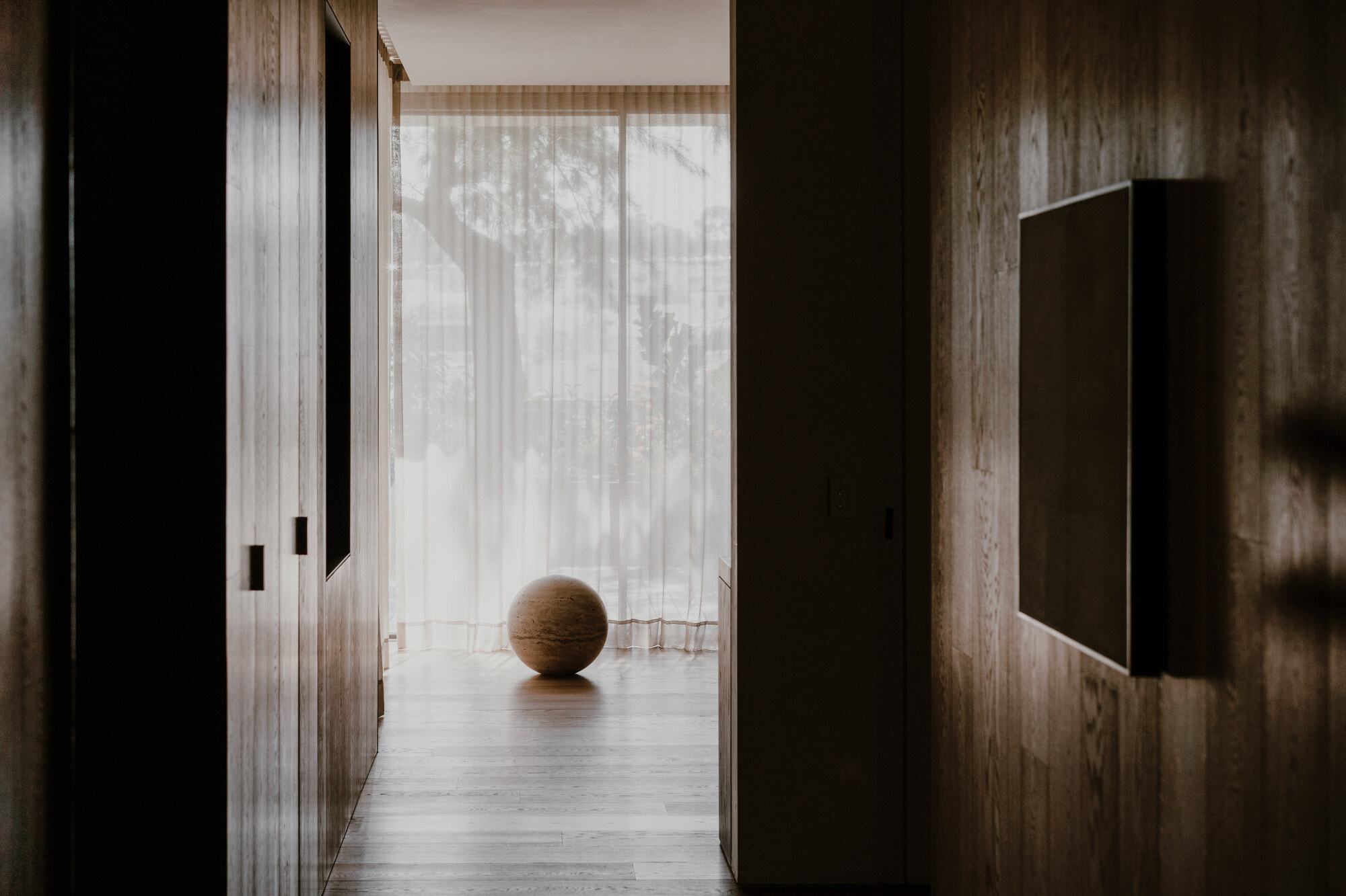Timber-paneled hallway framing a stone sphere on the floor in front of sheer curtains and soft daylight