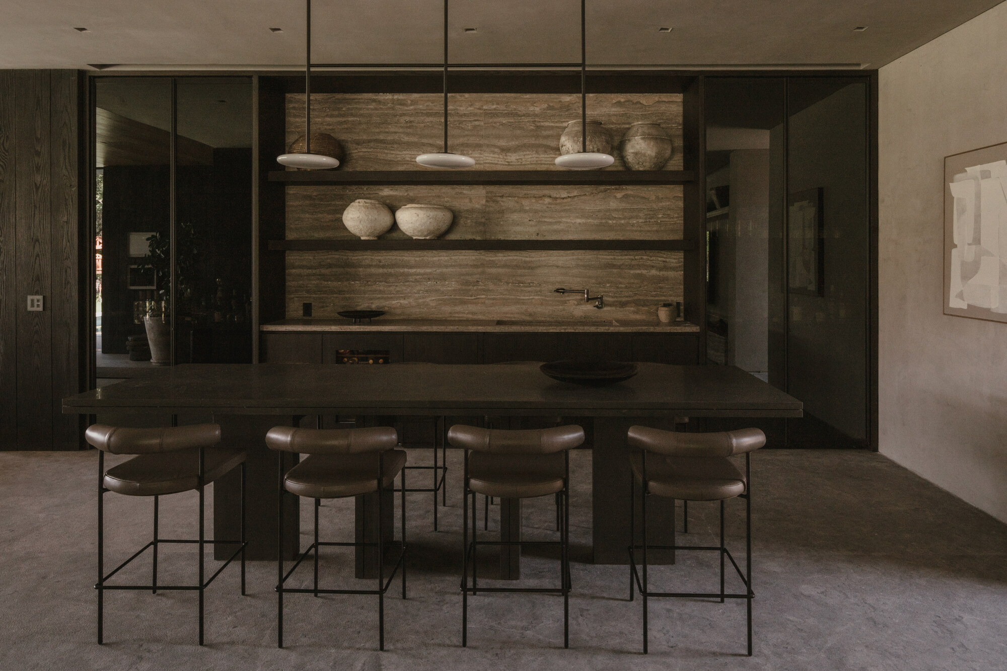 Kitchen wall of travertine shelving with pendant lamps and clay bowls behind a dark island and leather-topped stools