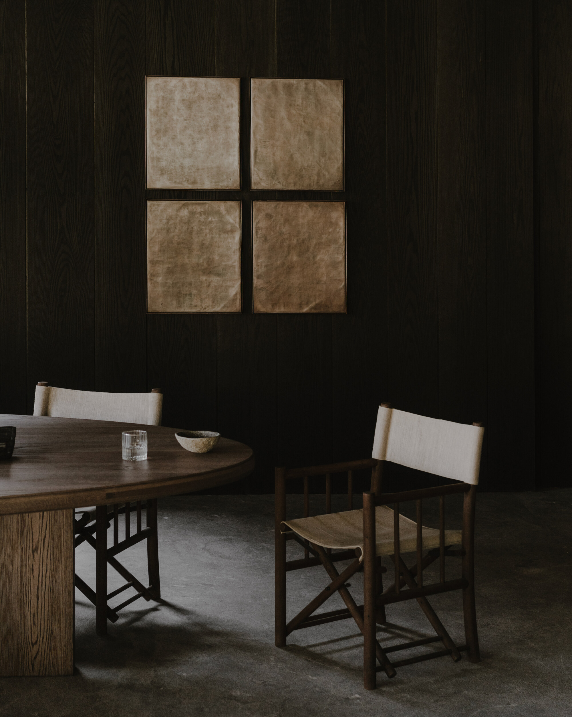 Four gilded paper panels on a dark wood wall above a round table and folding canvas-backed chairs