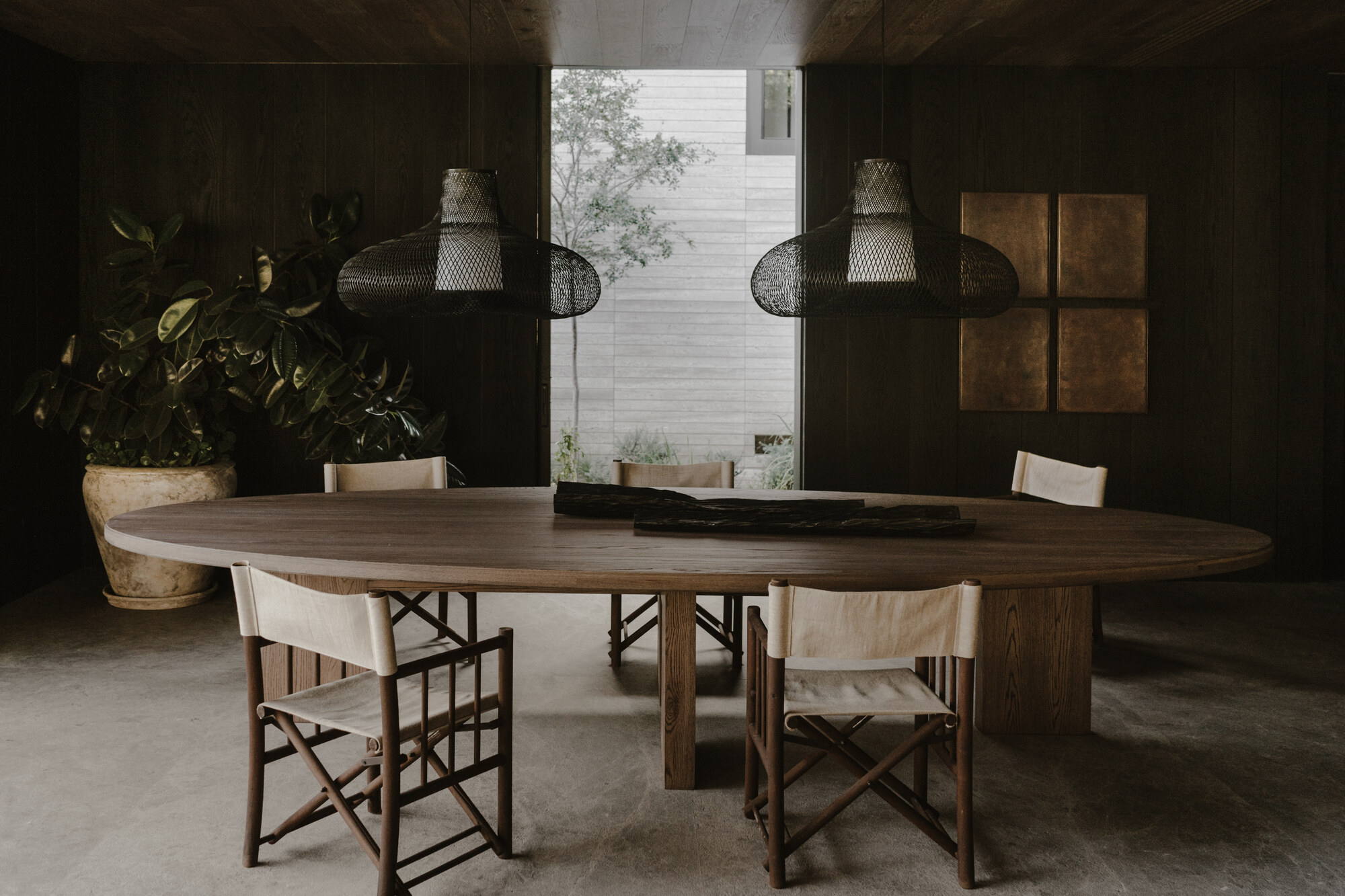 Oval dining table with folding chairs under woven black pendants, a potted rubber plant and a courtyard tree beyond the glass
