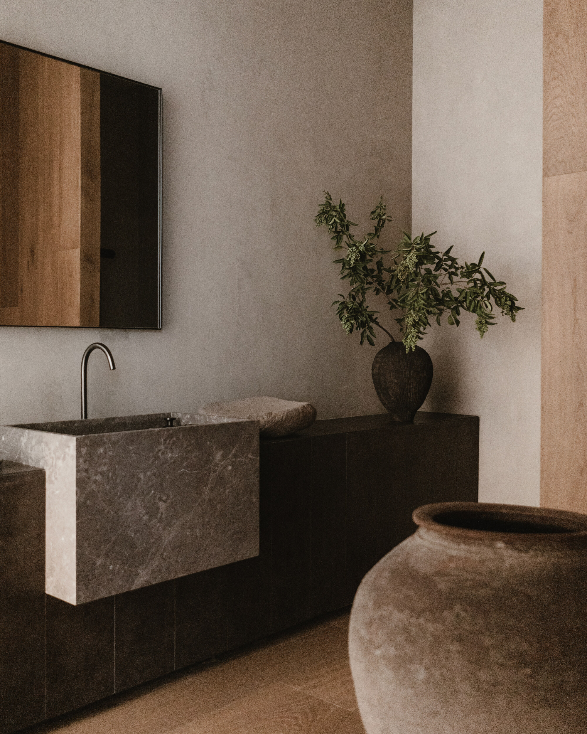 Stone basin with a slim arched tap on a dark wood vanity, with a clay jar and olive branches in soft light