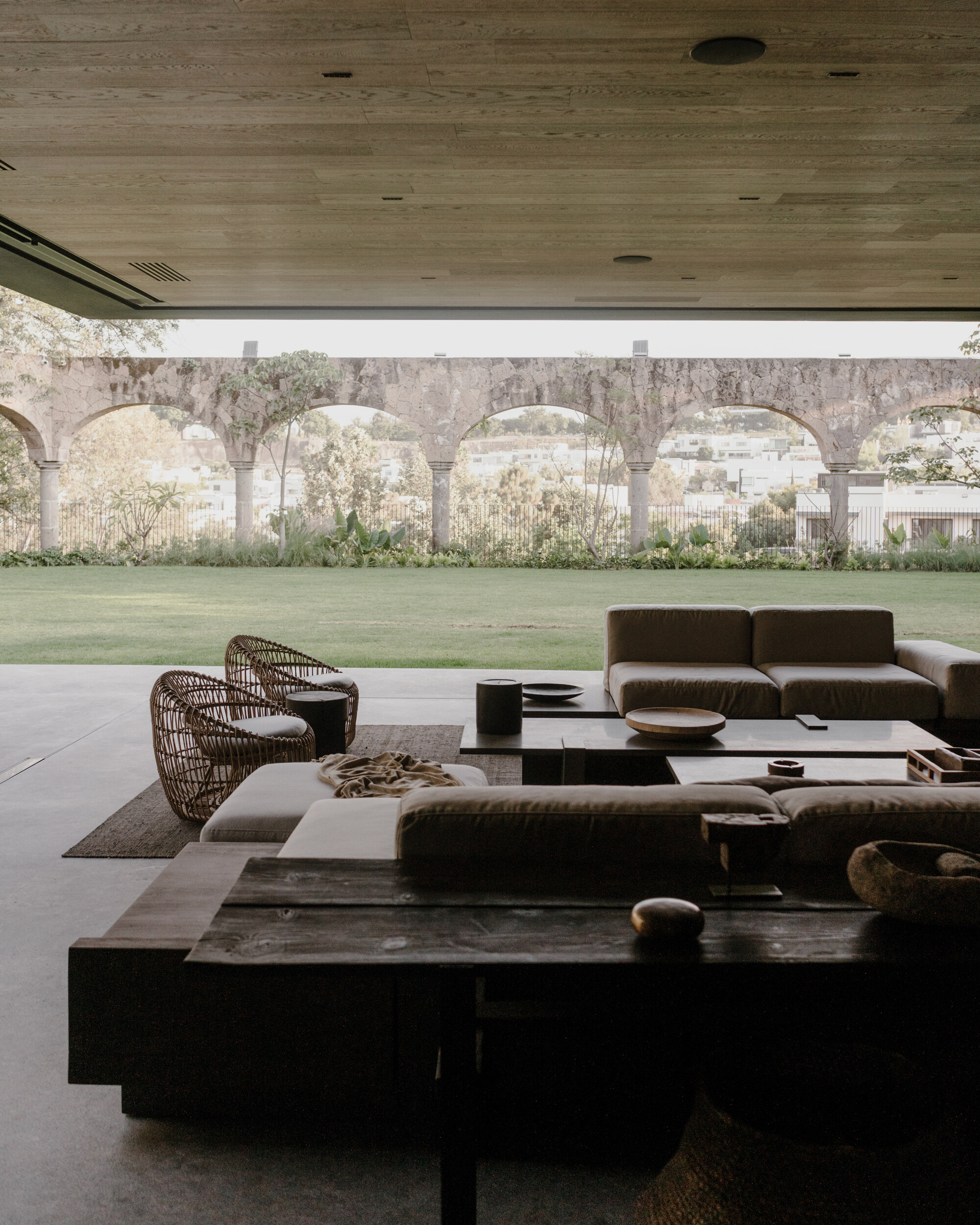Open living terrace with low sofas and wicker chairs facing a lawn and a stone aqueduct arcade under a timber ceiling