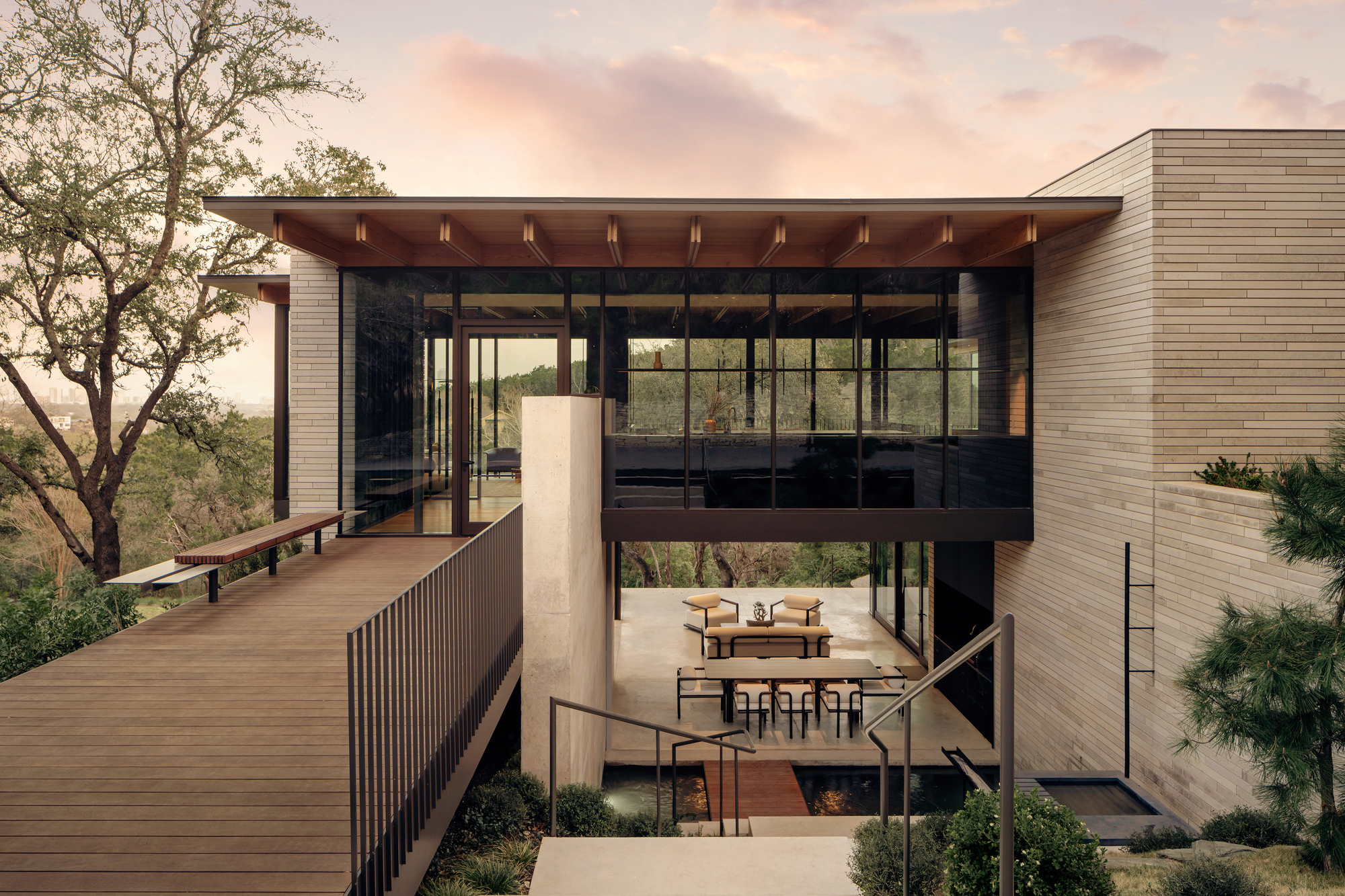Glass-walled upper volume with a timber soffit above a courtyard dining terrace, seen from a slatted deck at dusk