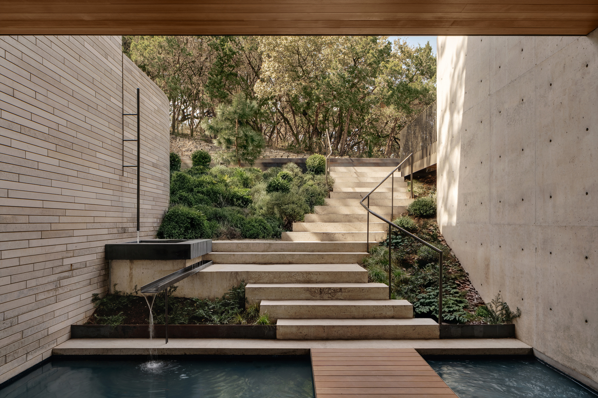 Concrete and stone steps rising between a board-formed wall and a brick wall, with a water spout feeding a pool below