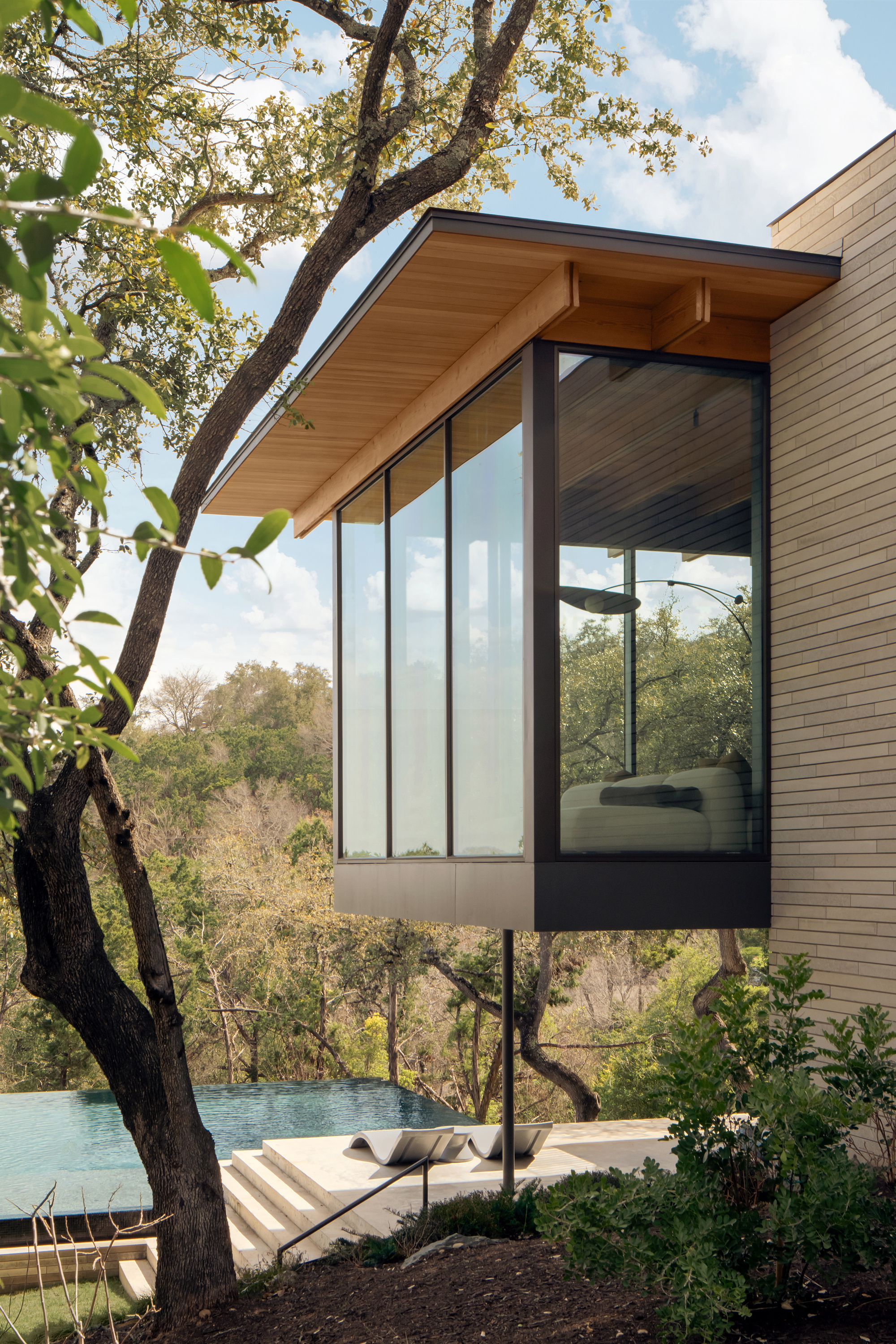 Corner bedroom box cantilevered on a steel post with a timber overhang, framed by an oak branch and a pool beyond