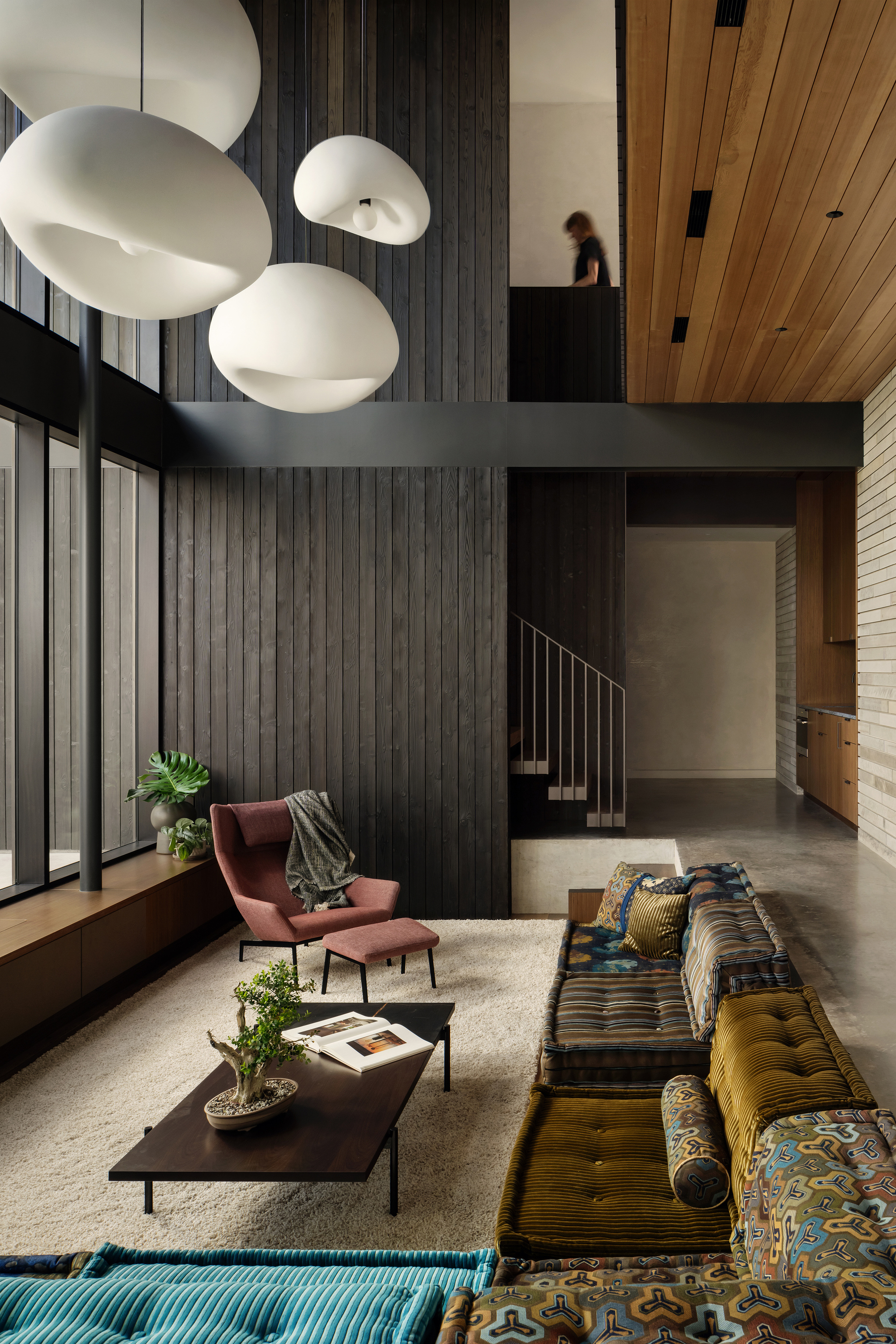 Double-height living room with white pendant lamps, charred timber wall, a mauve armchair and a patterned low sofa, a figure on the landing above